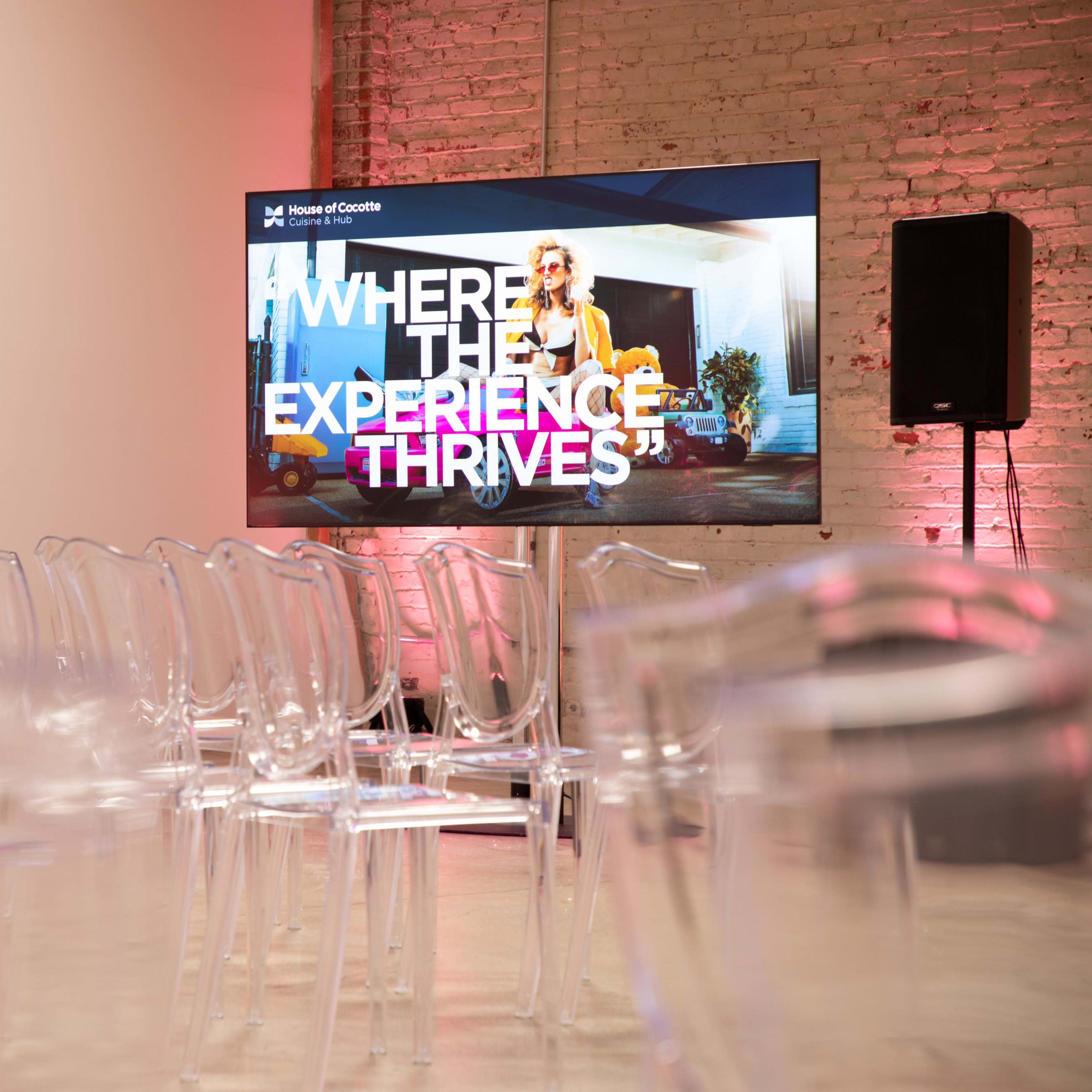 A modern event space with clear chairs arranged in rows facing a large screen that displays text saying, "Where the Experience Thrives."