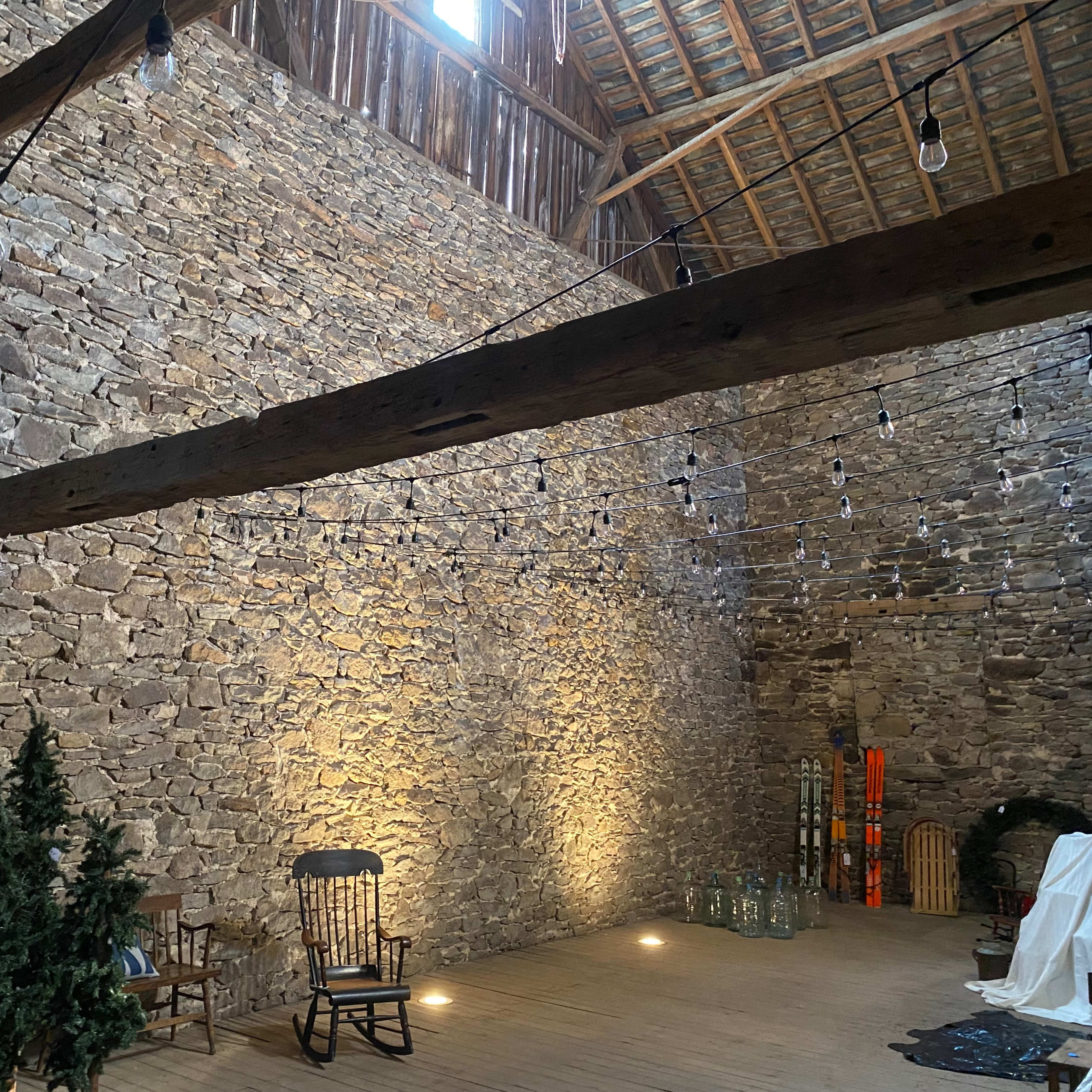 A rustic barn interior with a stone wall, wooden beams, and a wooden floor, decorated with string lights and a few pieces of furniture.