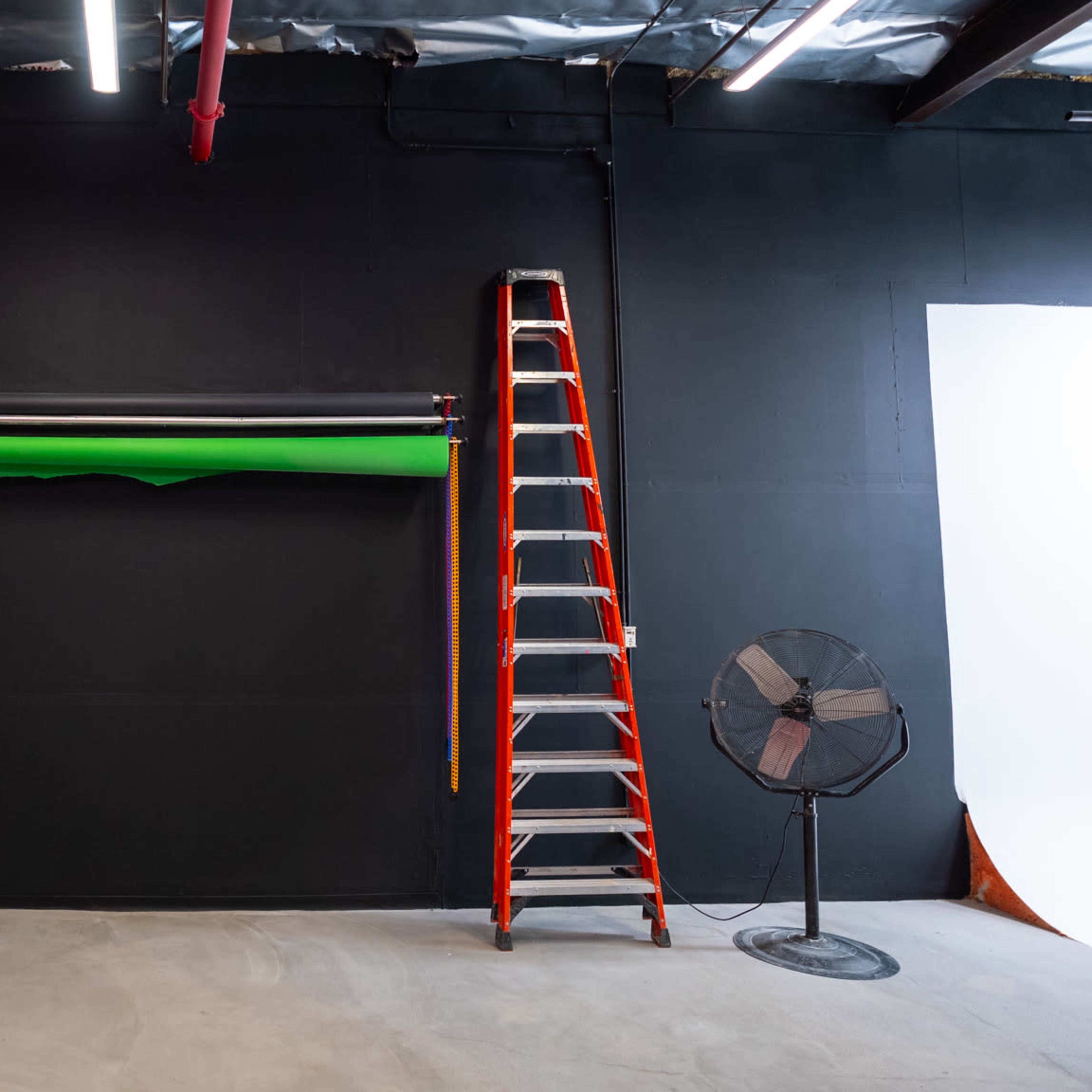 A black-walled studio features a green backdrop roll, a ladder, trash bins, a broom, and a fan.