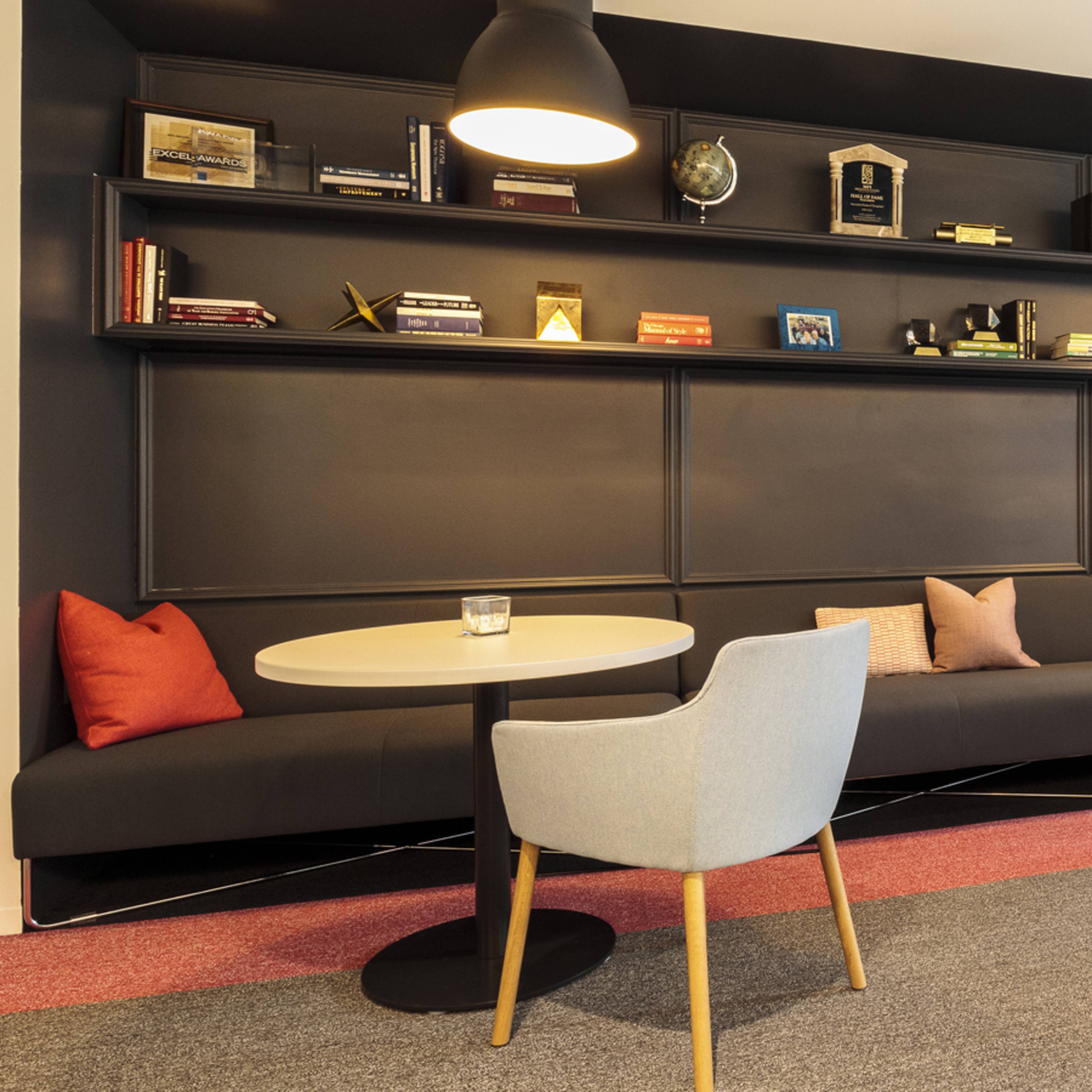 A modern lounge area with a dark shelving unit displaying various items, two round tables, and comfortable seating with pillows.