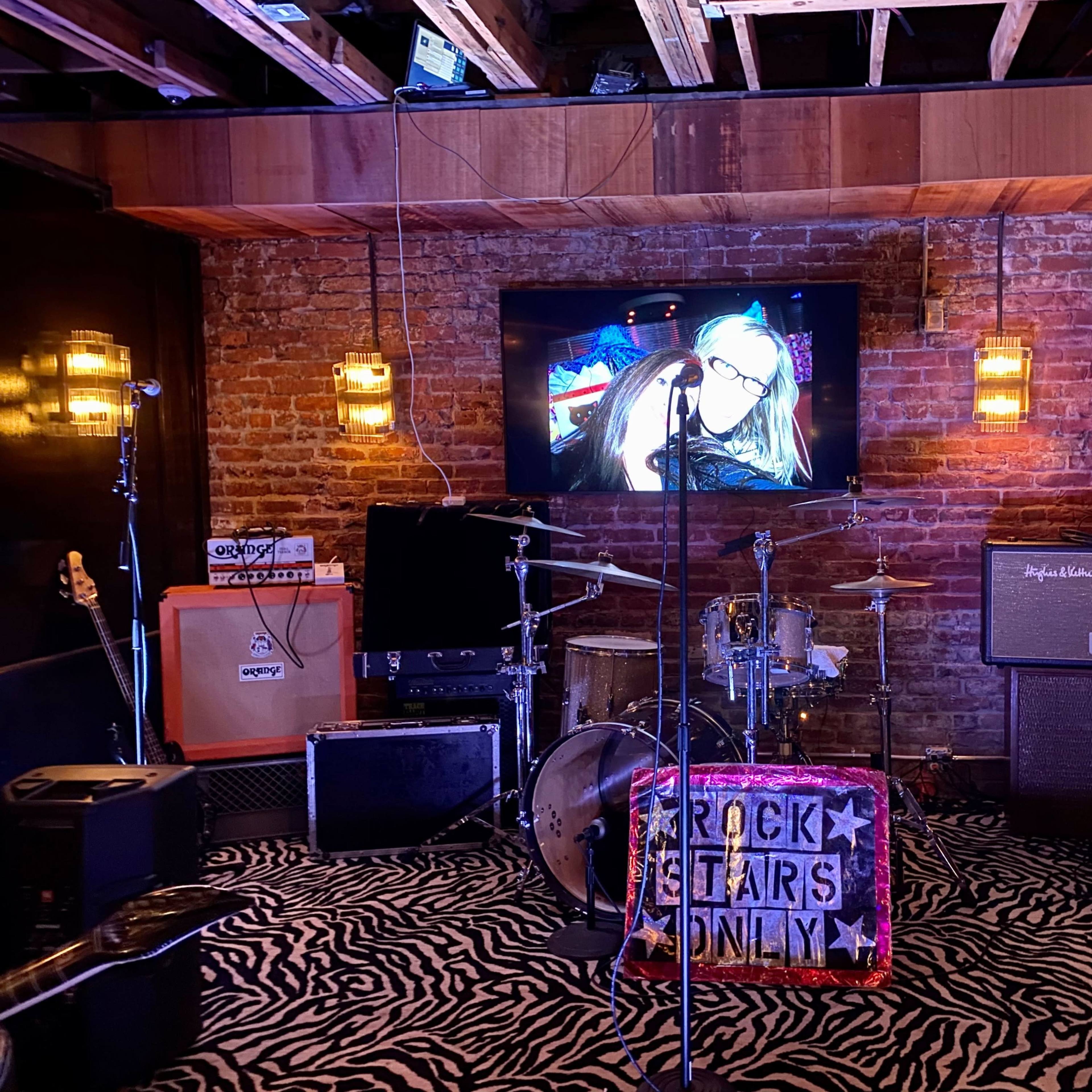 A stage set up for a live music performance features various instruments, amplifiers, and a screen displaying an image on a rustic brick wall.