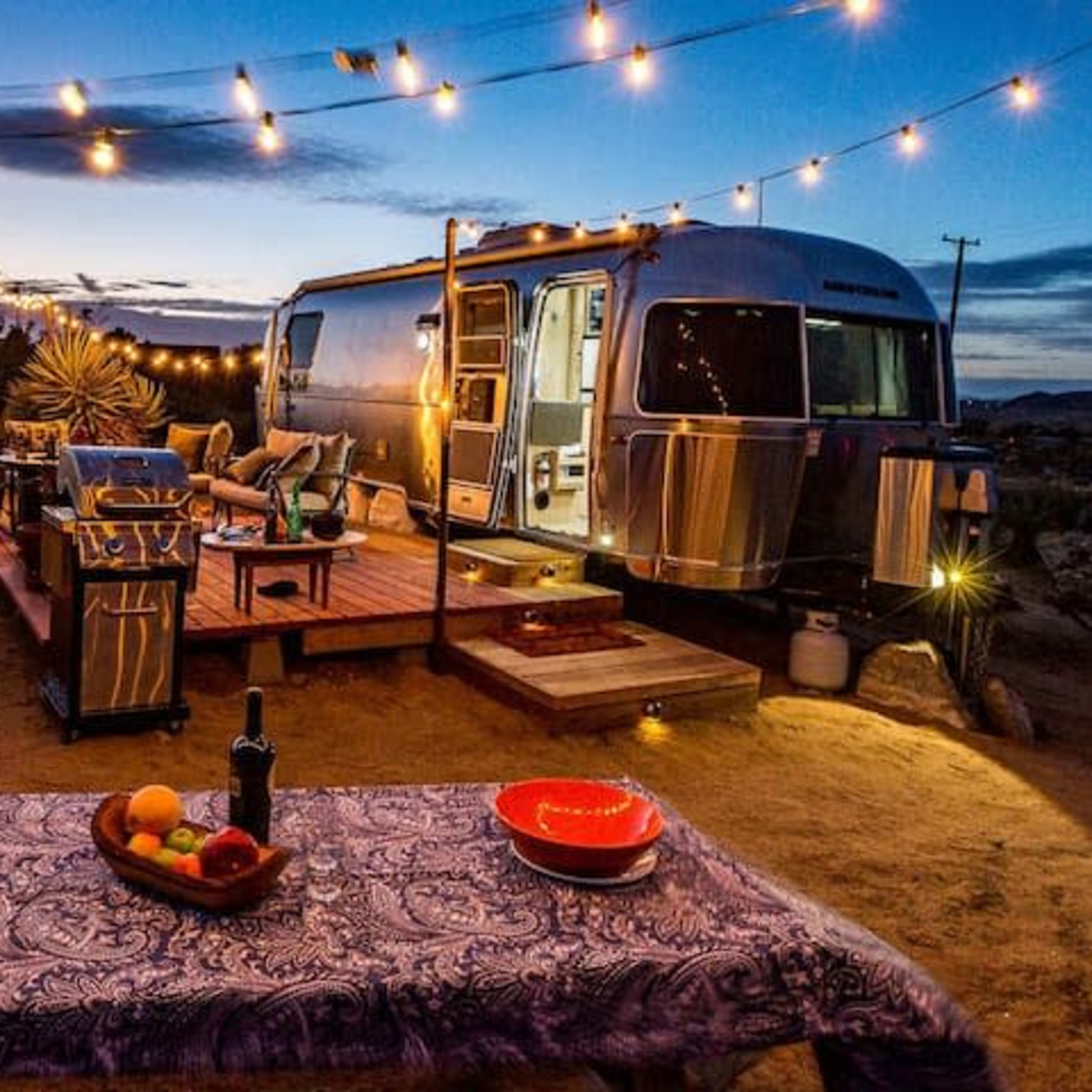 A retro Airstream trailer is parked on a desert lot, illuminated by string lights and surrounded by outdoor furniture and a table set with fruit.