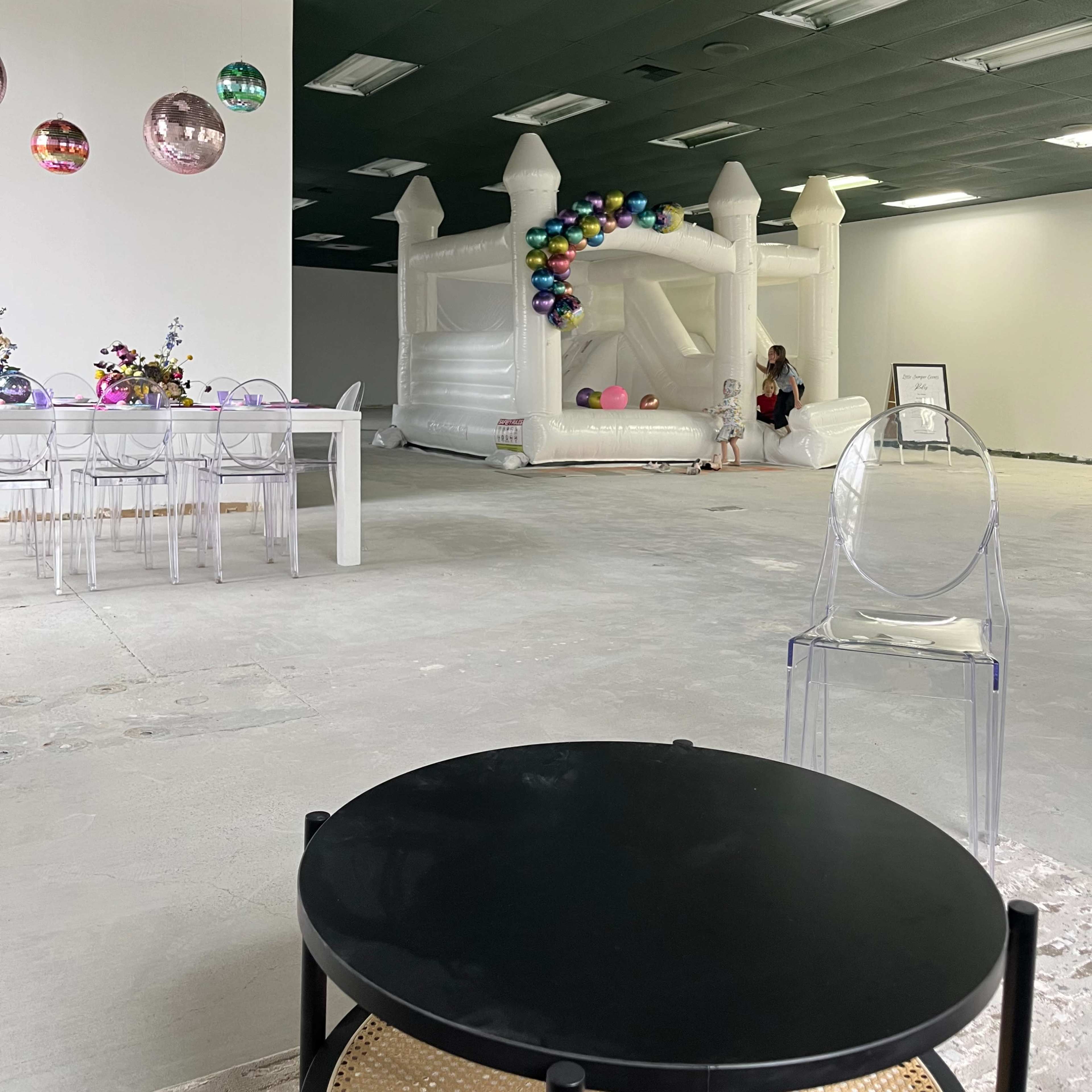 A bright event space featuring a colorful inflatable bounce house, a table set for dining, and transparent chairs arranged nearby.