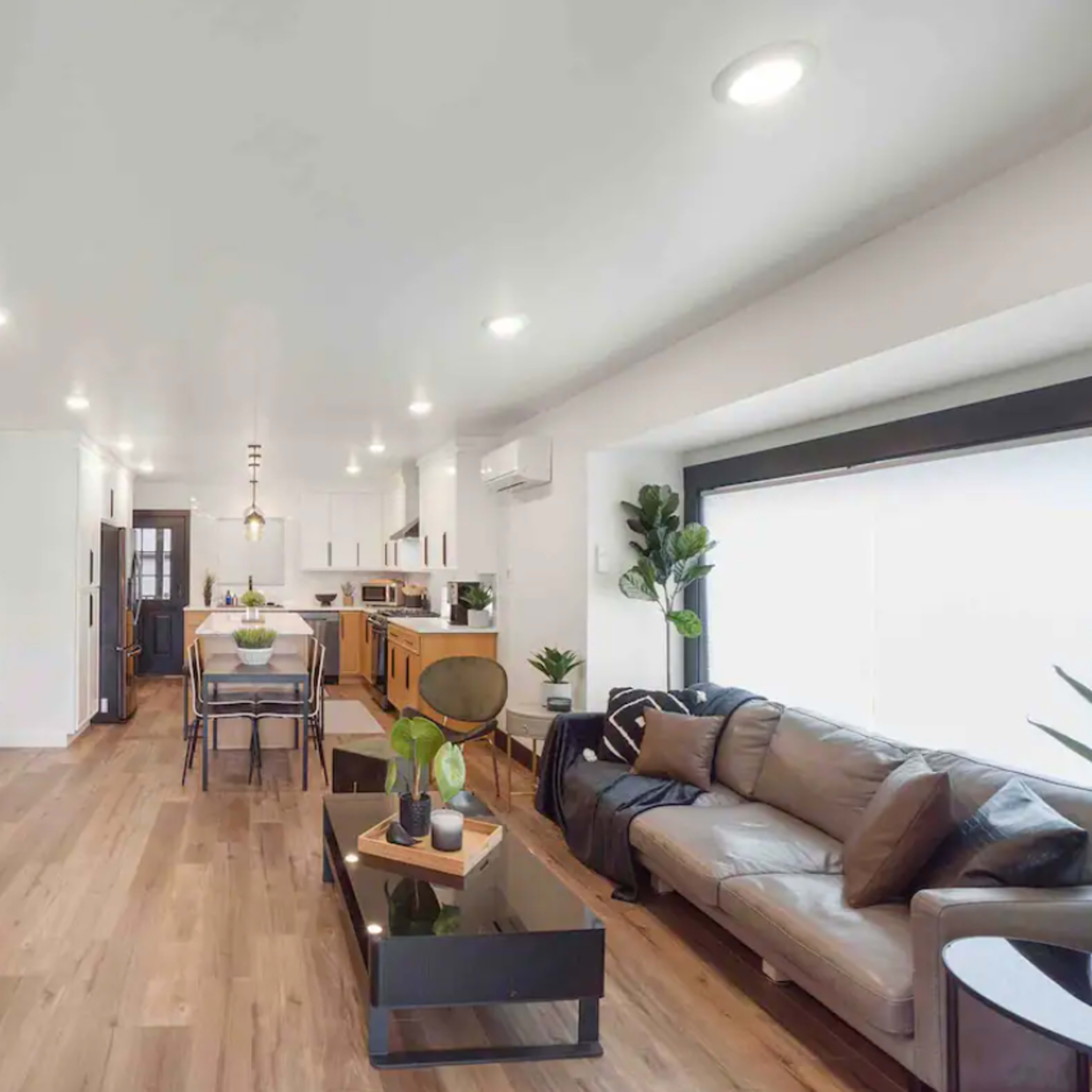 A modern living room with a large television, a coffee table, and a couch, leading into an open kitchen area with dining space.