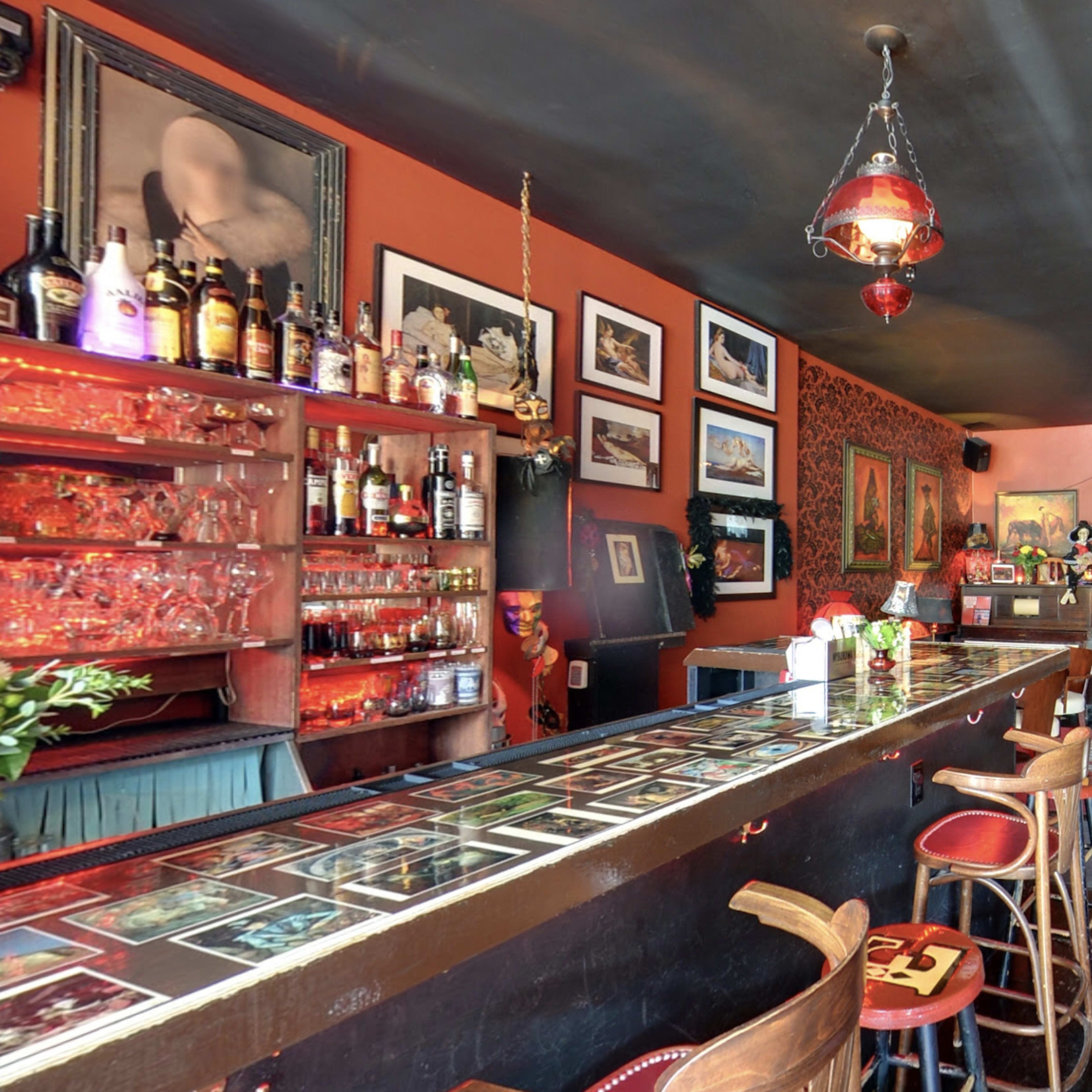 A dimly lit bar features a long counter with a variety of alcohol bottles and several framed photographs on the walls.