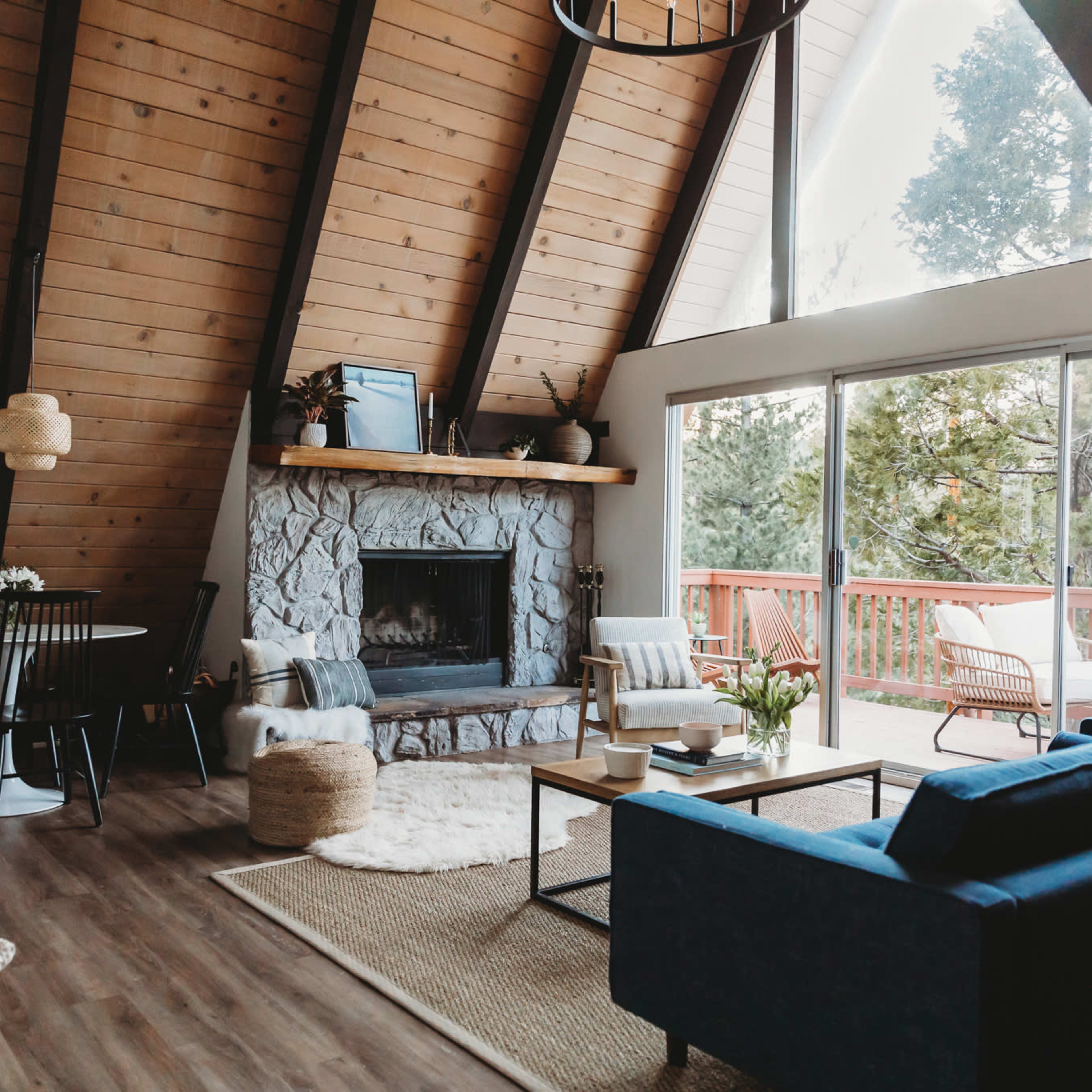 A modern A-frame living room with a stone fireplace, wooden beams, and large windows overlooking a deck and trees.