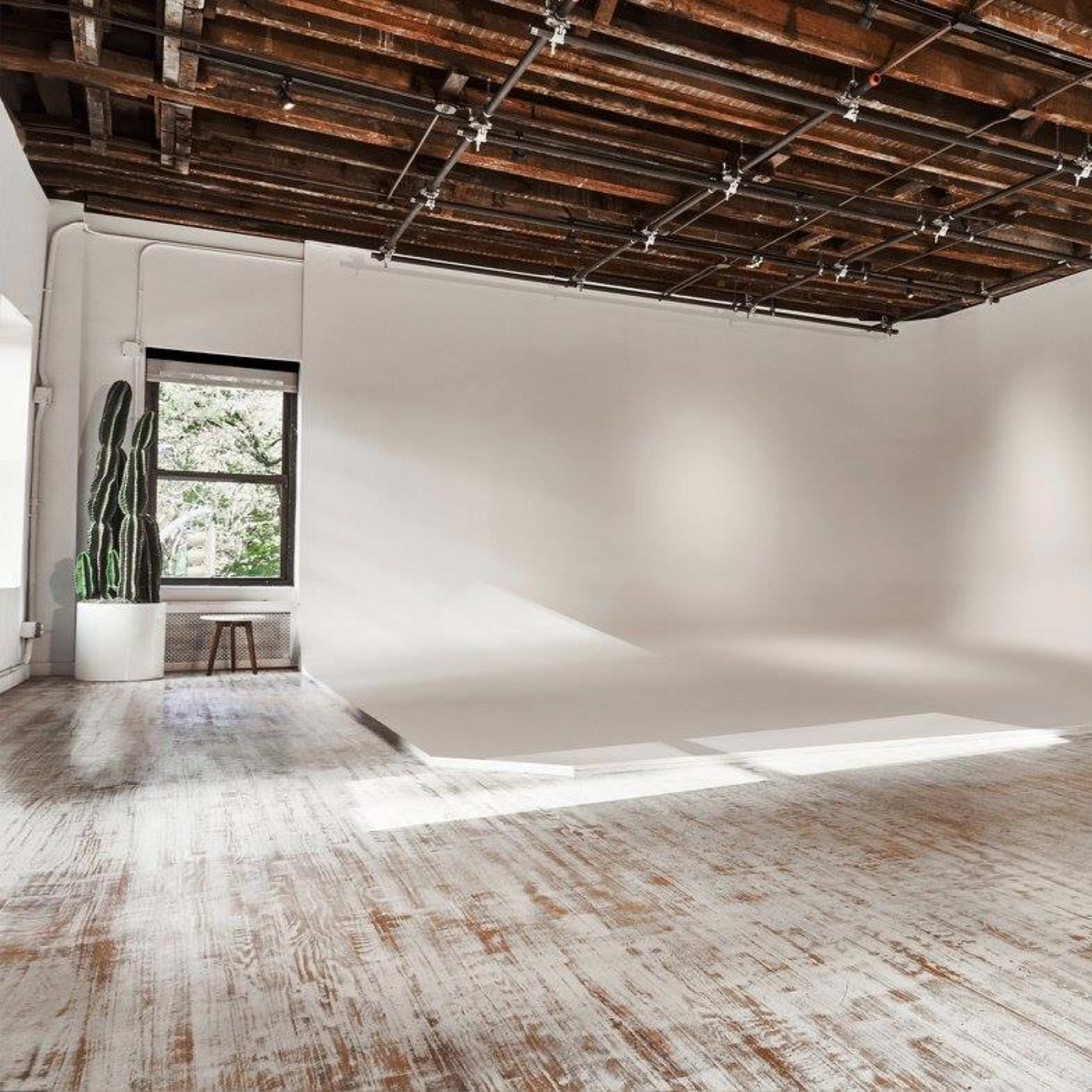 A spacious, empty studio with a white backdrop, wooden floor, and a large window letting in natural light.