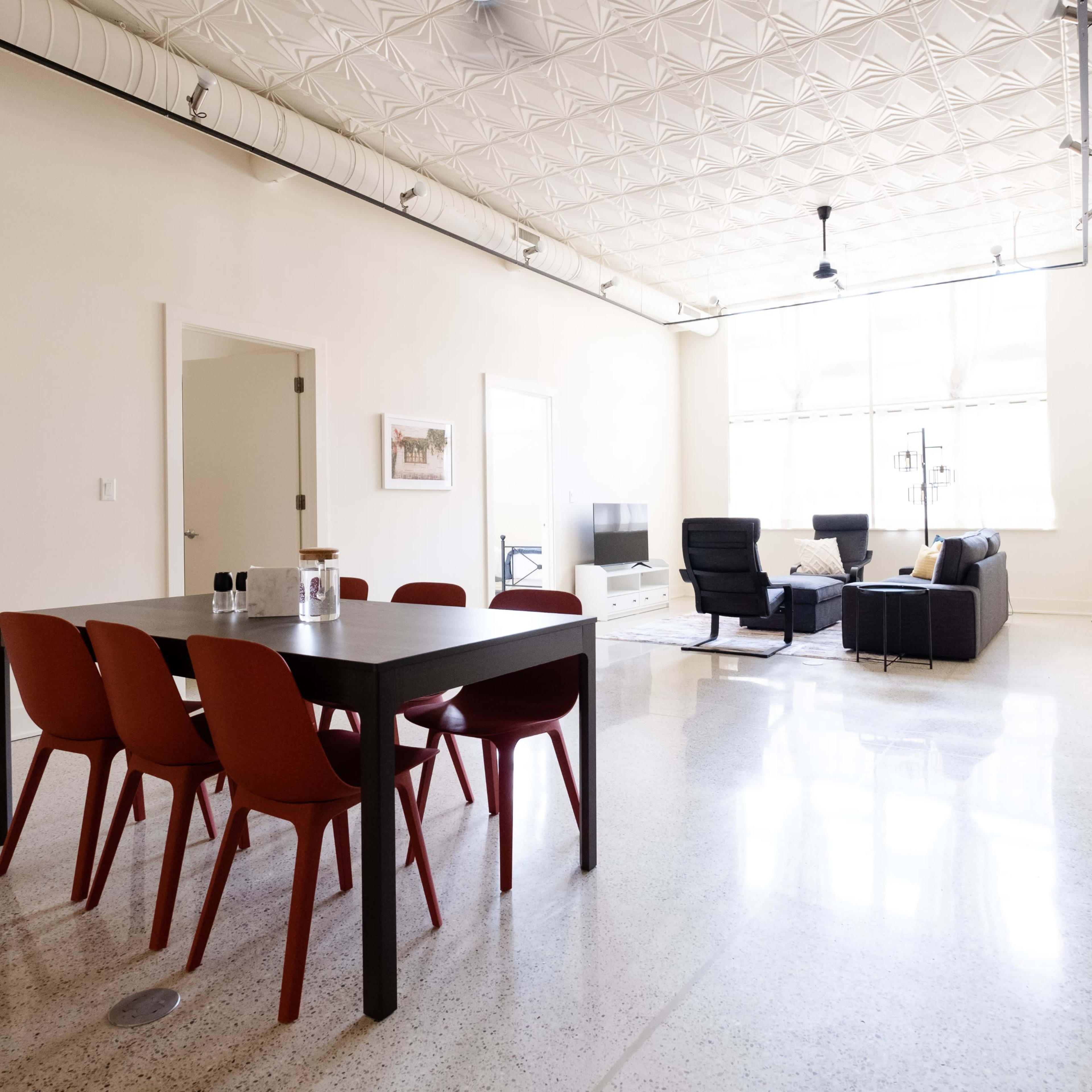 A dining table with six red chairs is situated in a spacious, brightly lit room featuring modern furnishings and a polished floor.