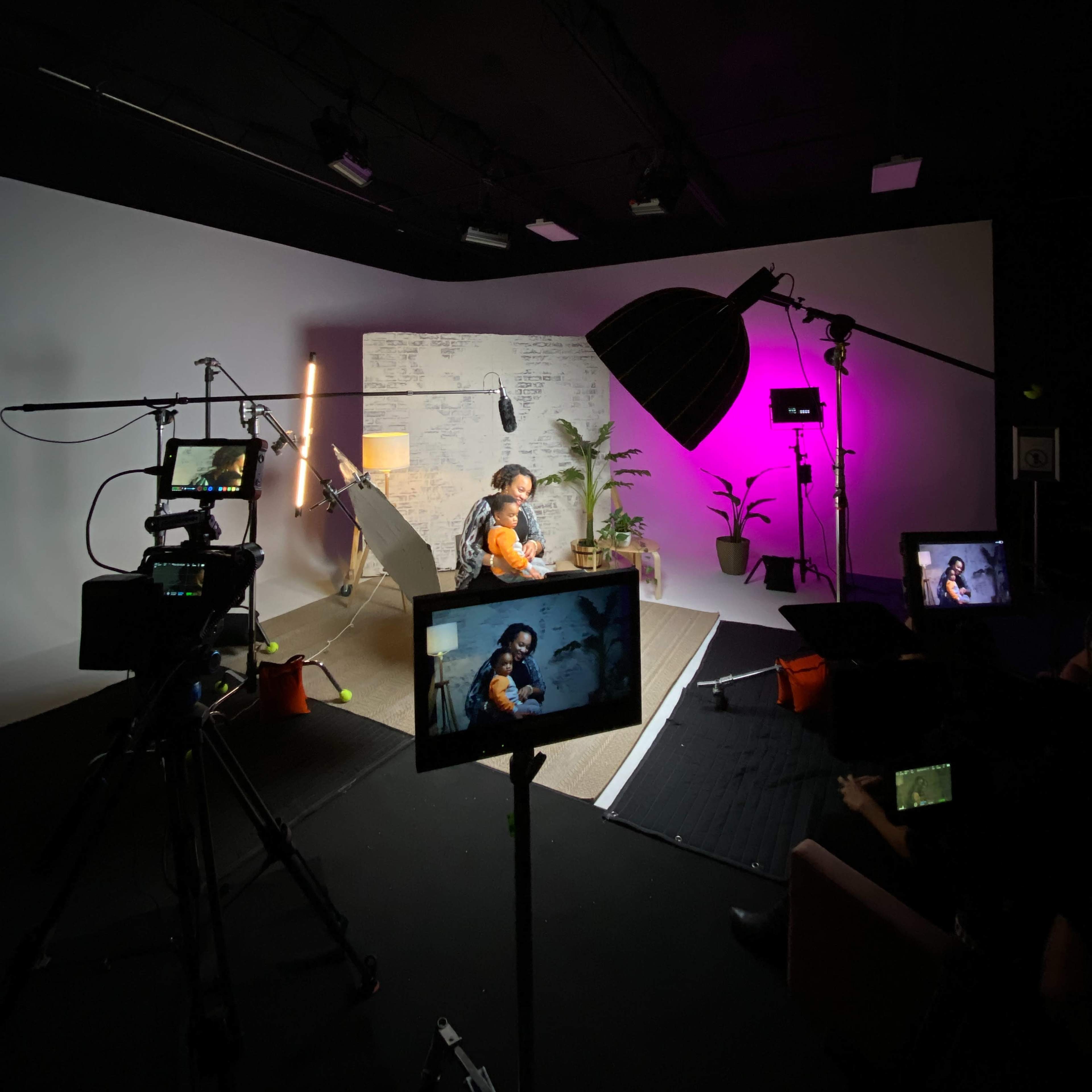 A studio setup is shown with a person seated against a backdrop, surrounded by cameras, lighting equipment, and potted plants.