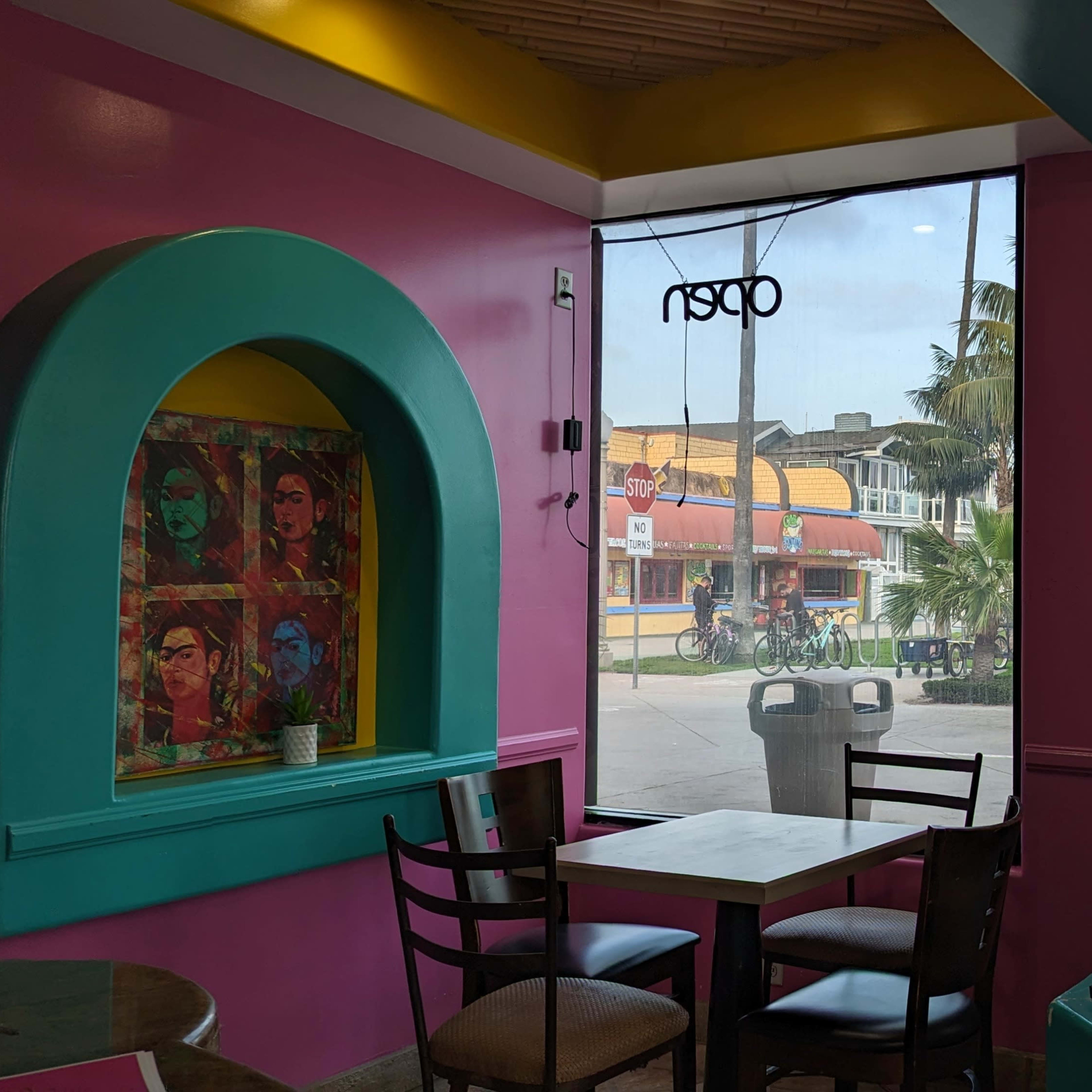 A colorful cafe interior with a view of a palm-lined street and shops through large glass doors.