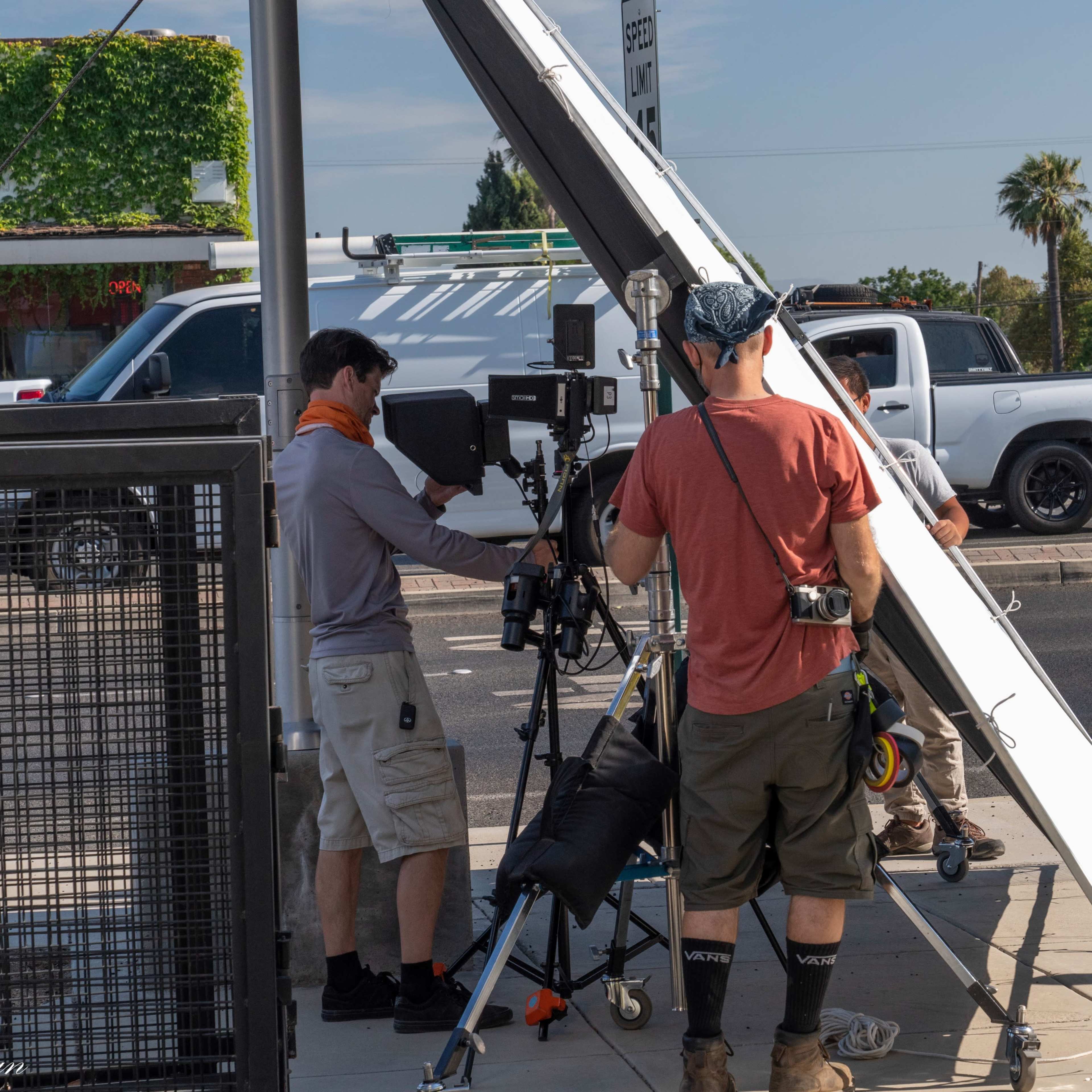 A film crew sets up equipment on a sidewalk near a restaurant and palm trees under clear skies.