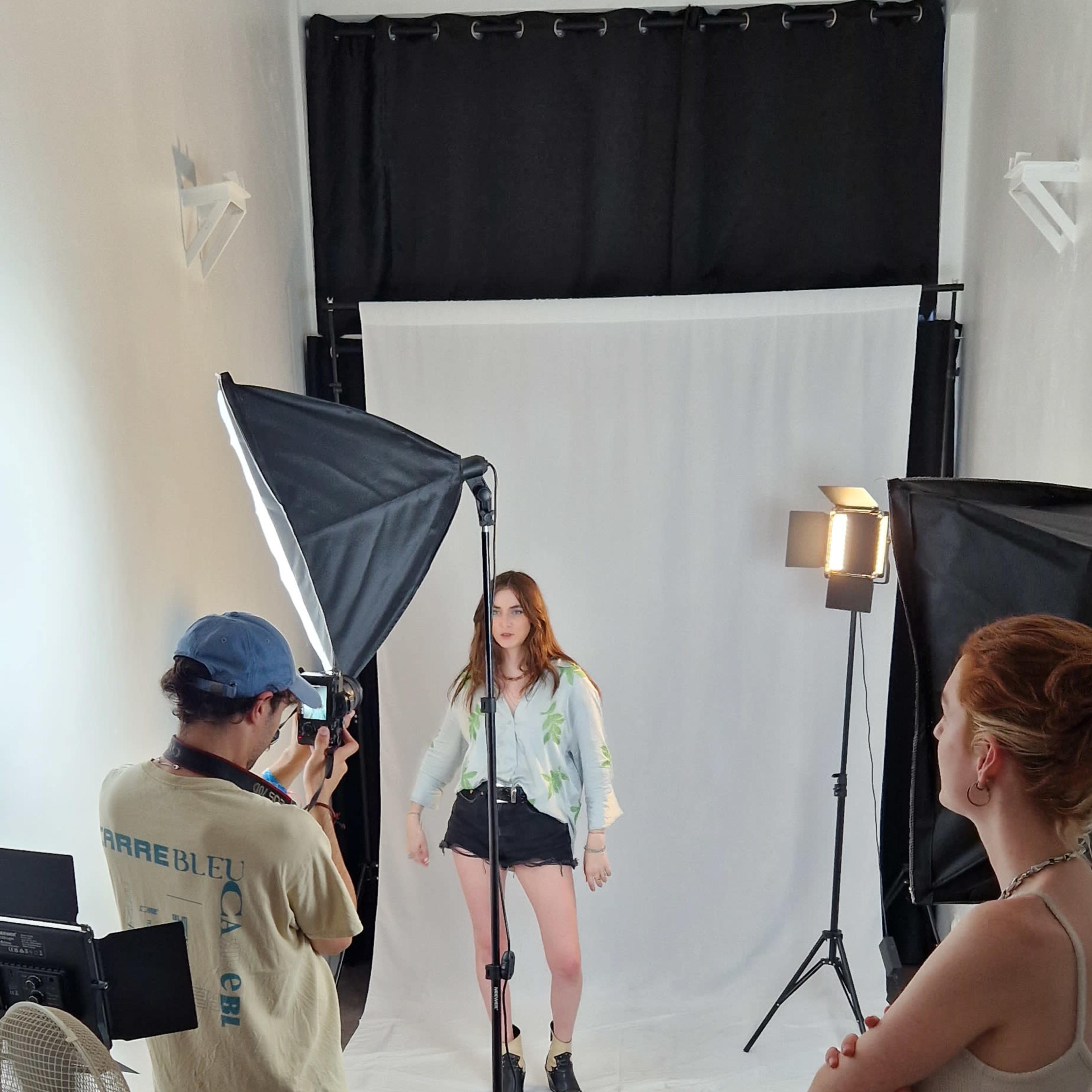 A model poses in front of a backdrop while a photographer captures the shot, surrounded by studio lights and a small crew.