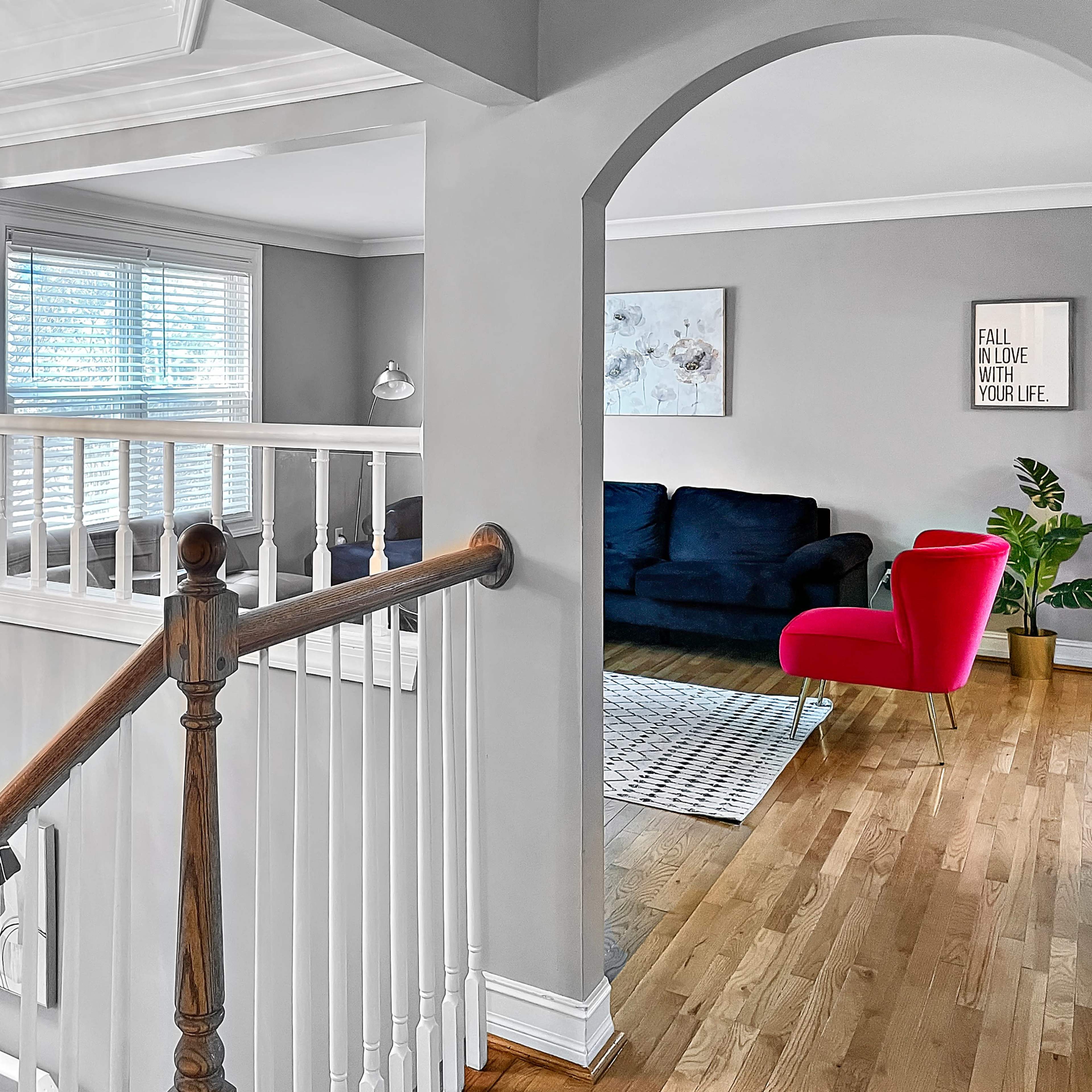 A staircase leading to a well-lit upper floor with a view of a living room featuring a dark sofa and a red accent chair.