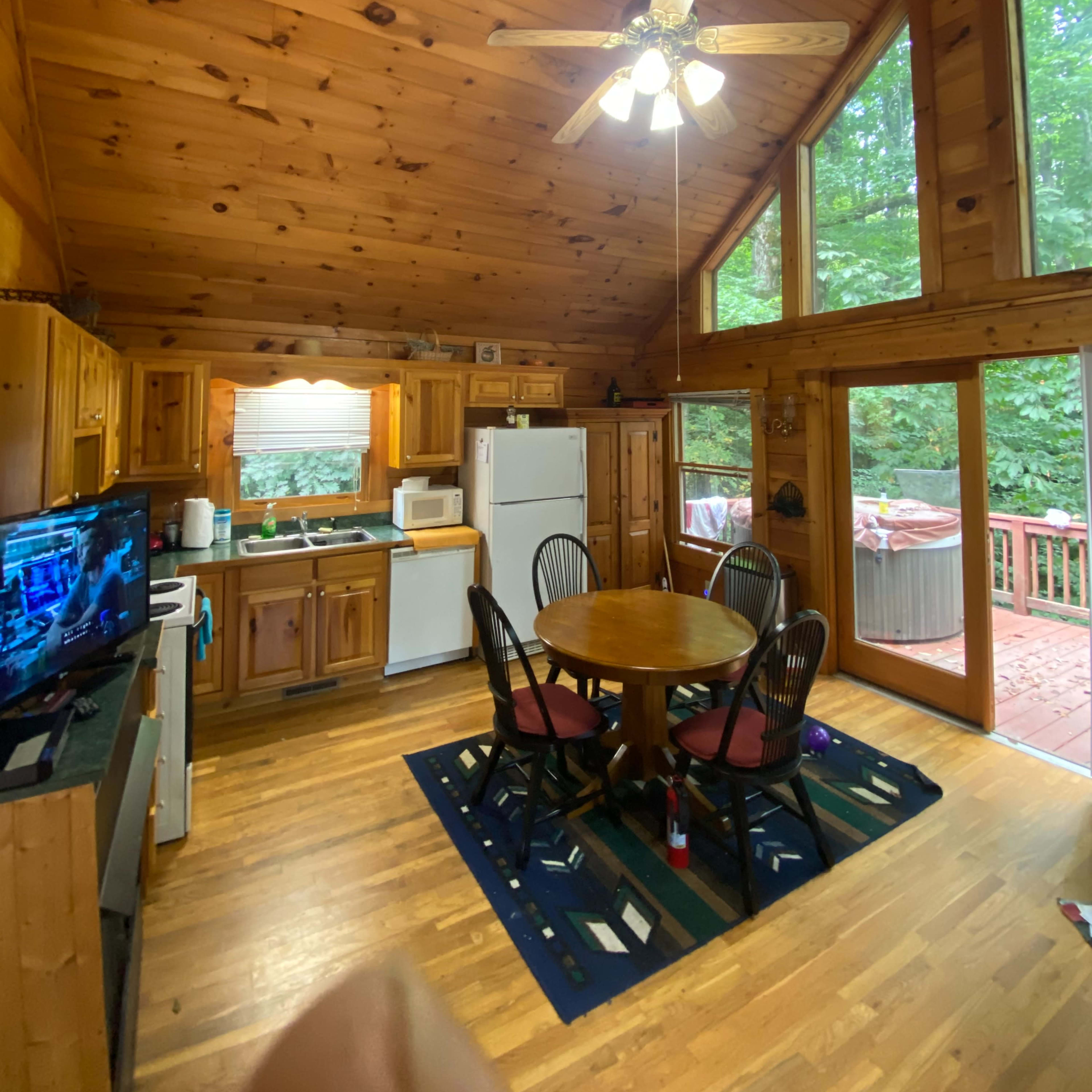 A wooden cabin kitchen with a circular dining table, four chairs, and large windows overlooking a deck.