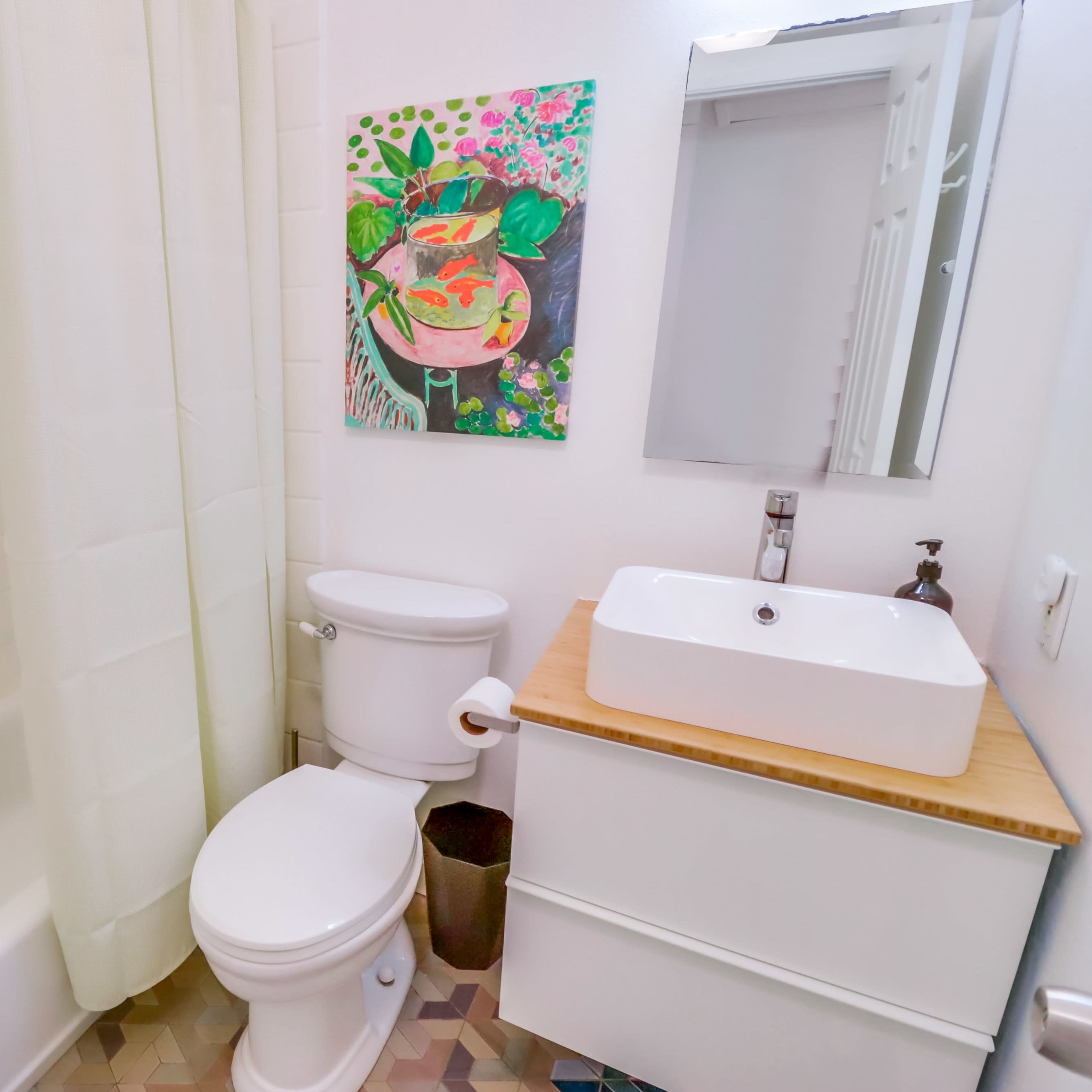 The bathroom features a bathtub with a shower curtain, a toilet, a wall-mounted sink, and a colorful painting above the sink.