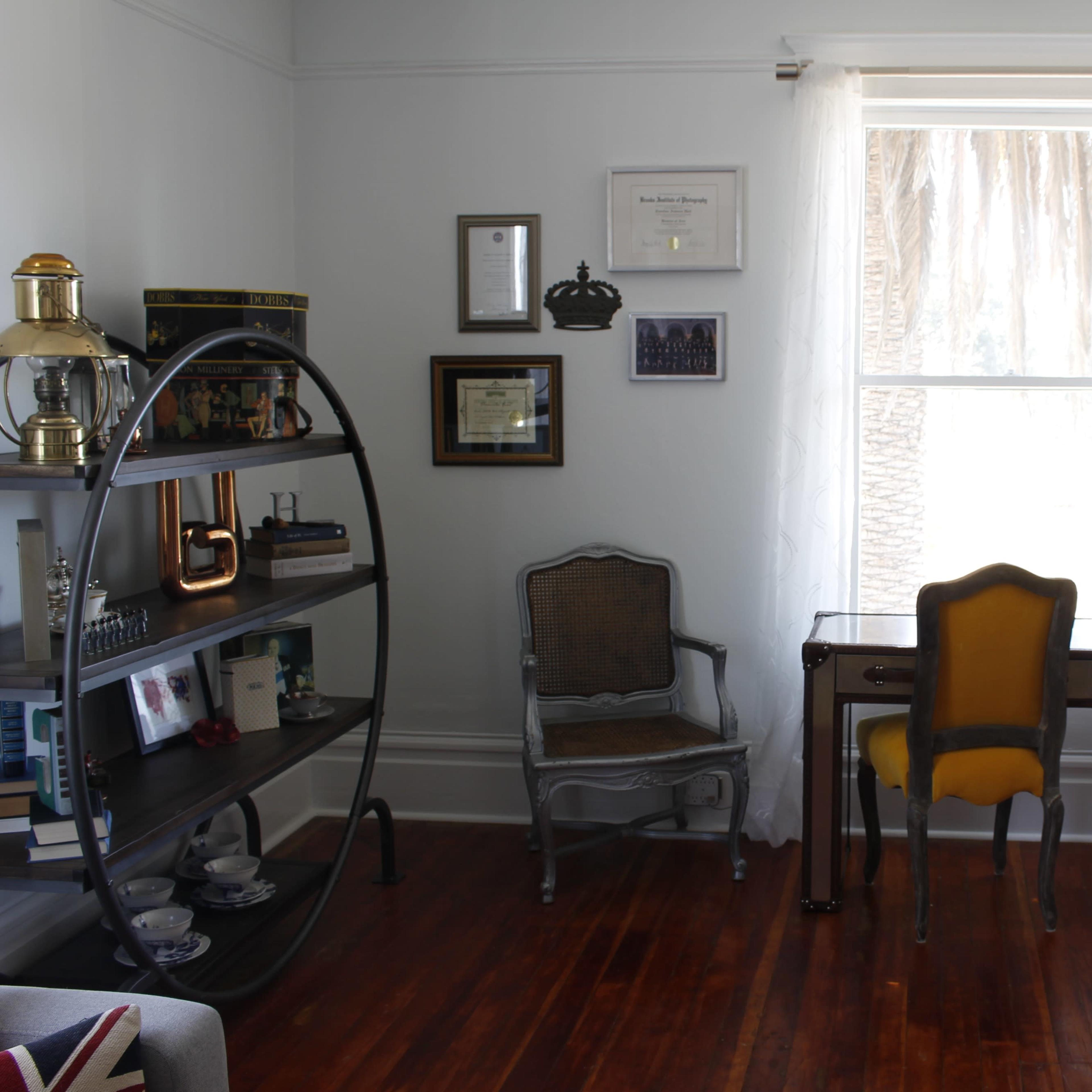 The room features a small desk with a yellow chair, a bookshelf with various items, and a window draped with sheer curtains.