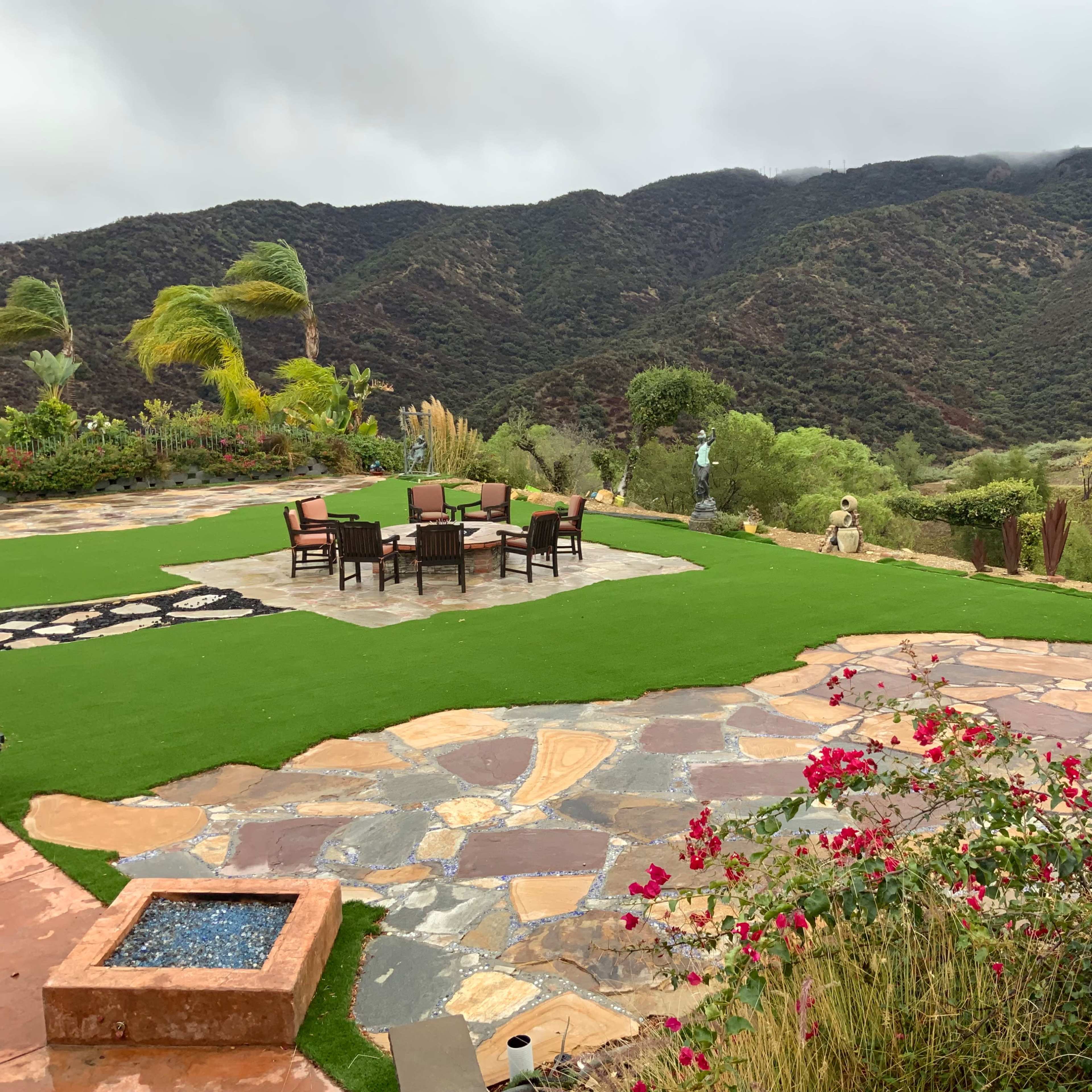 A landscaped outdoor area with a stone patio, a seating arrangement around a table, and a mountainous backdrop.