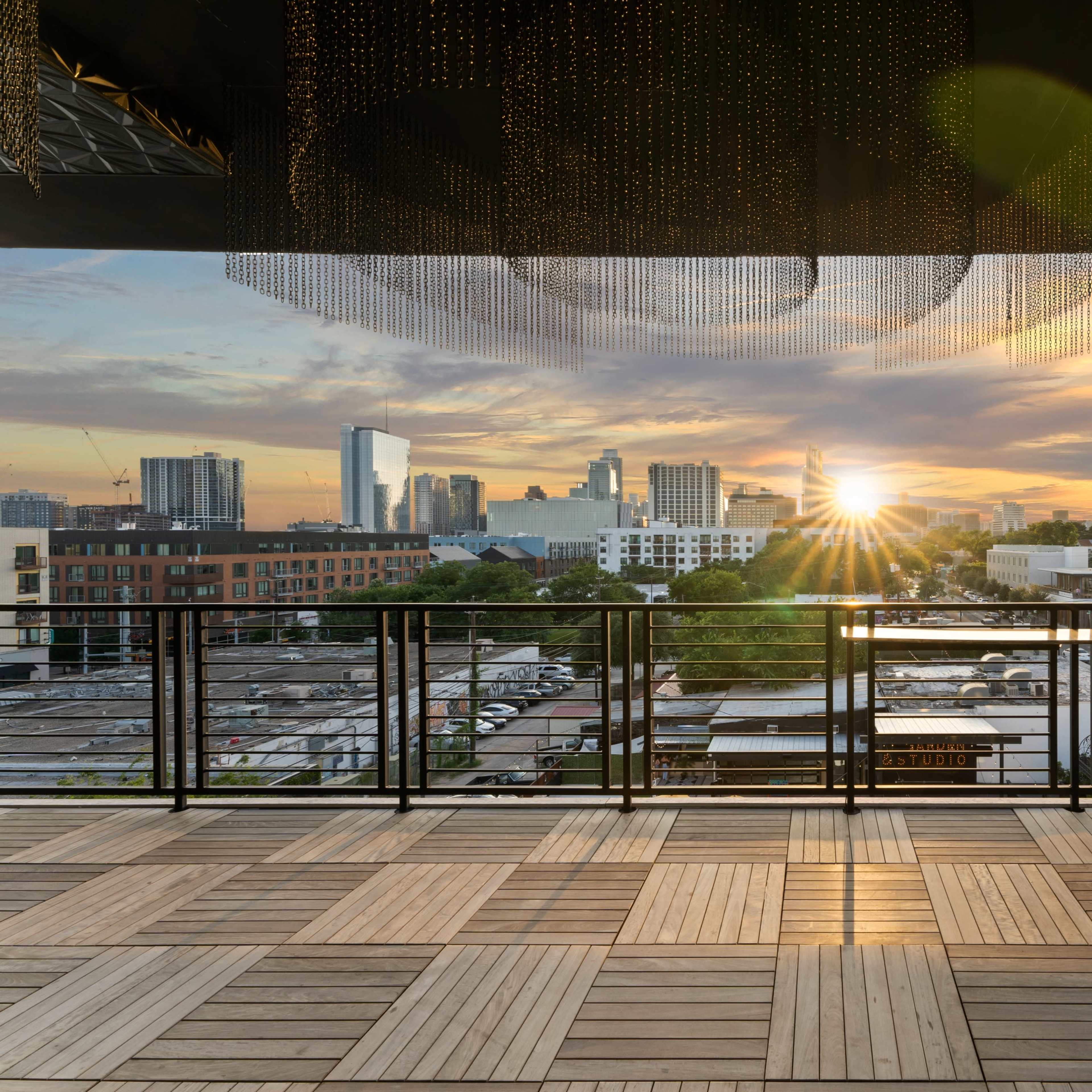 A balcony with a wooden floor overlooking a city skyline at sunset, with the sun illuminating the buildings.