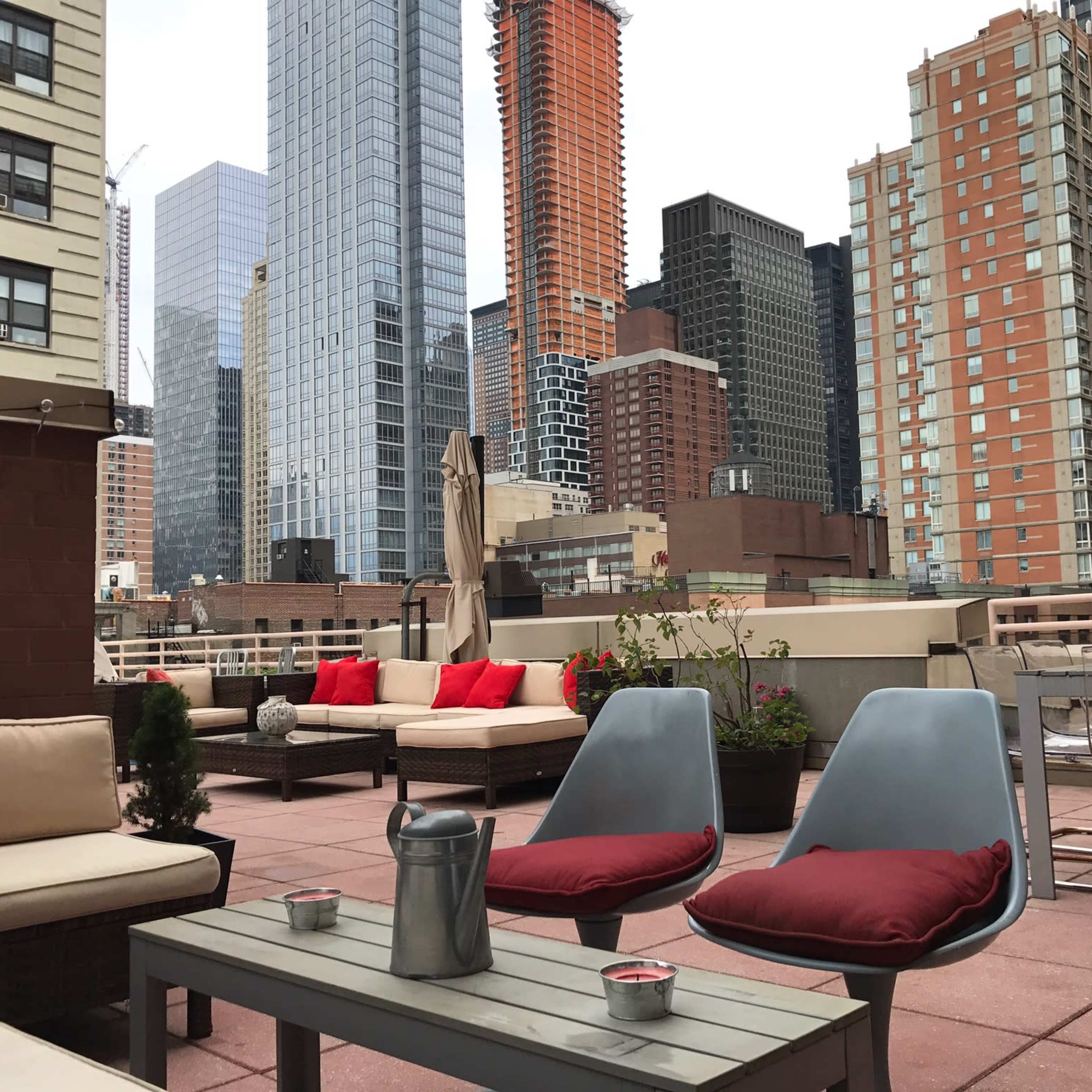 A rooftop terrace features seating arrangements with cushions, landscaped planters, and a skyline of tall buildings in the background.