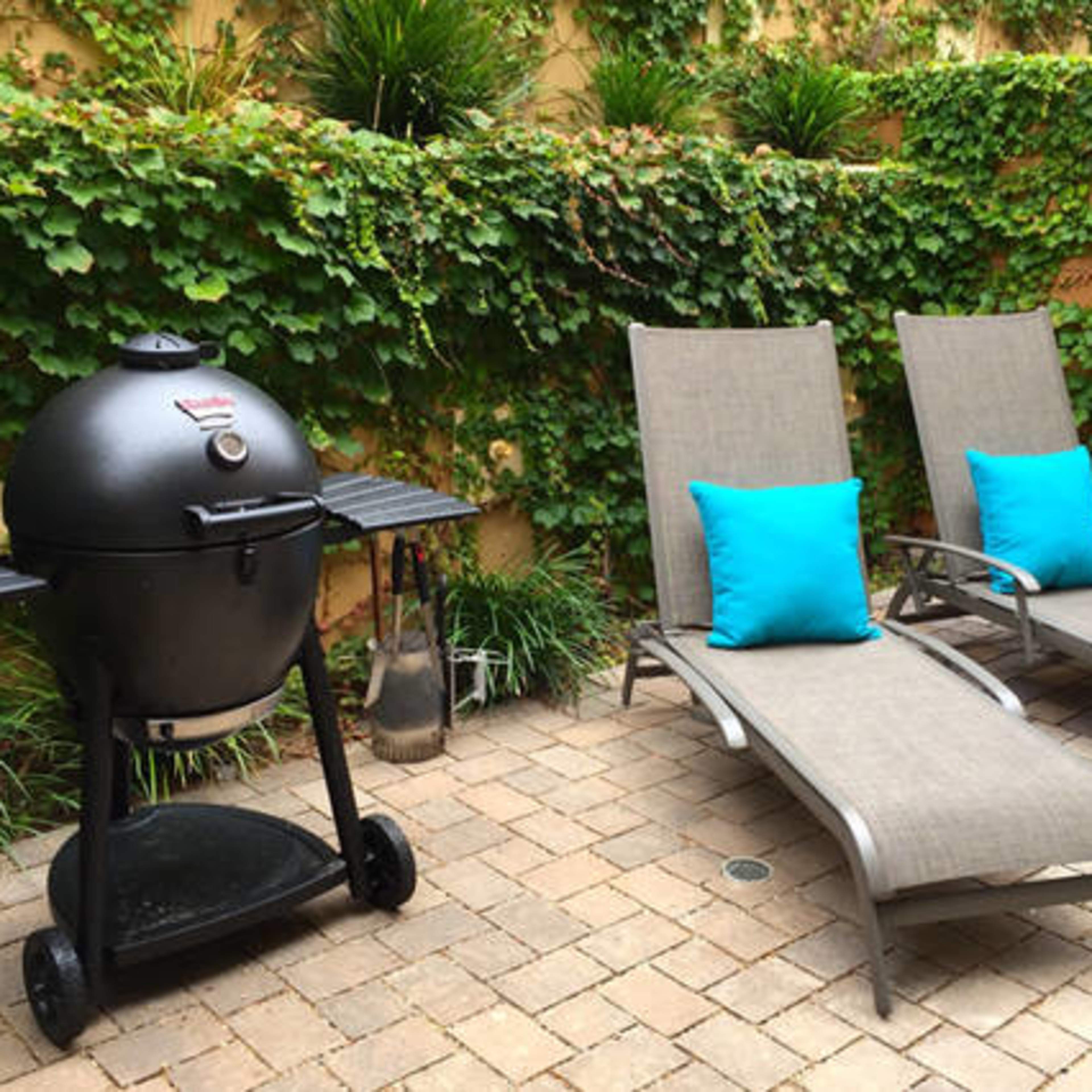 A black grill next to two gray lounge chairs with turquoise cushions, set in a landscaped outdoor area with leafy greenery.