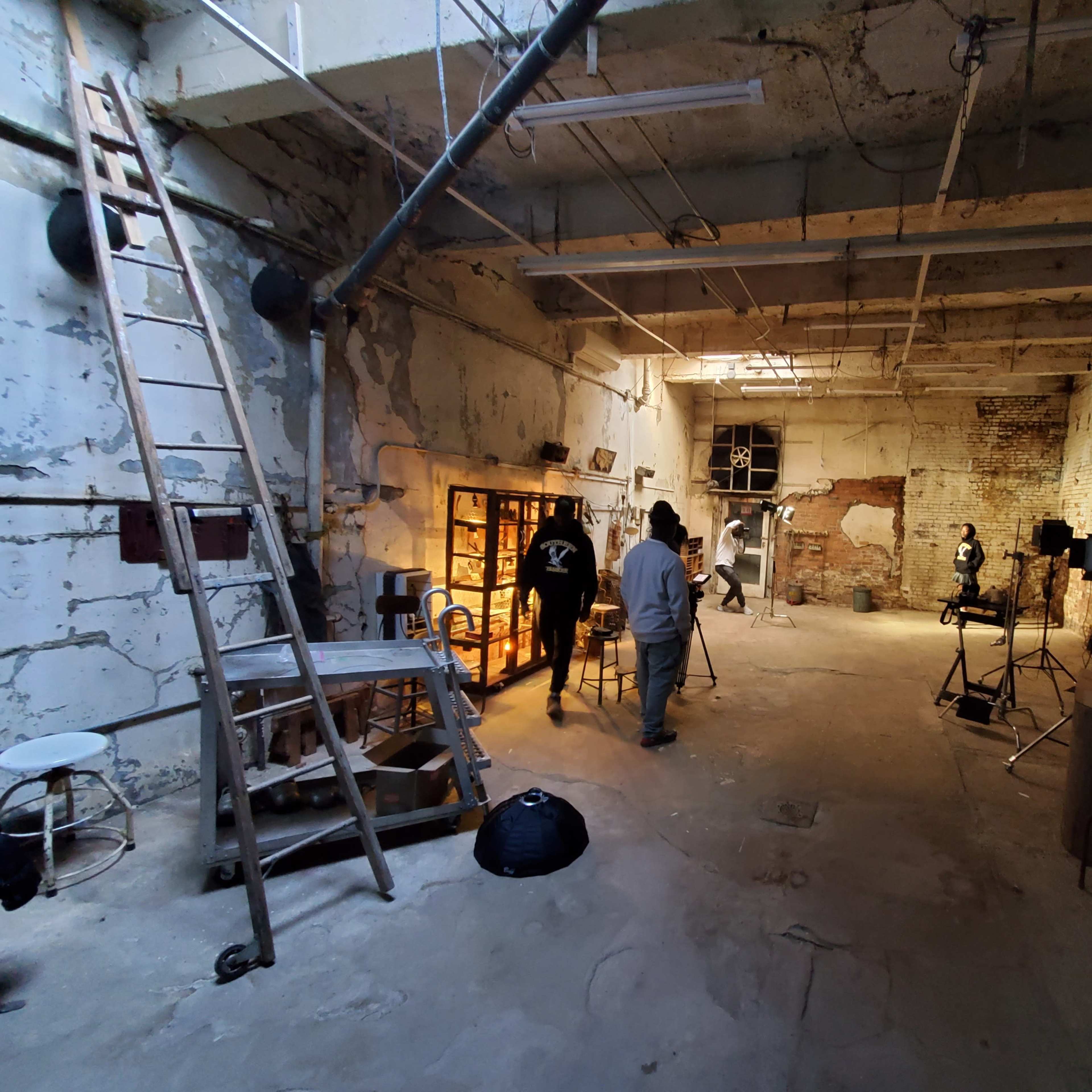An industrial space with exposed brick walls, a concrete floor, and several individuals setting up equipment amidst various furniture and lighting setups.