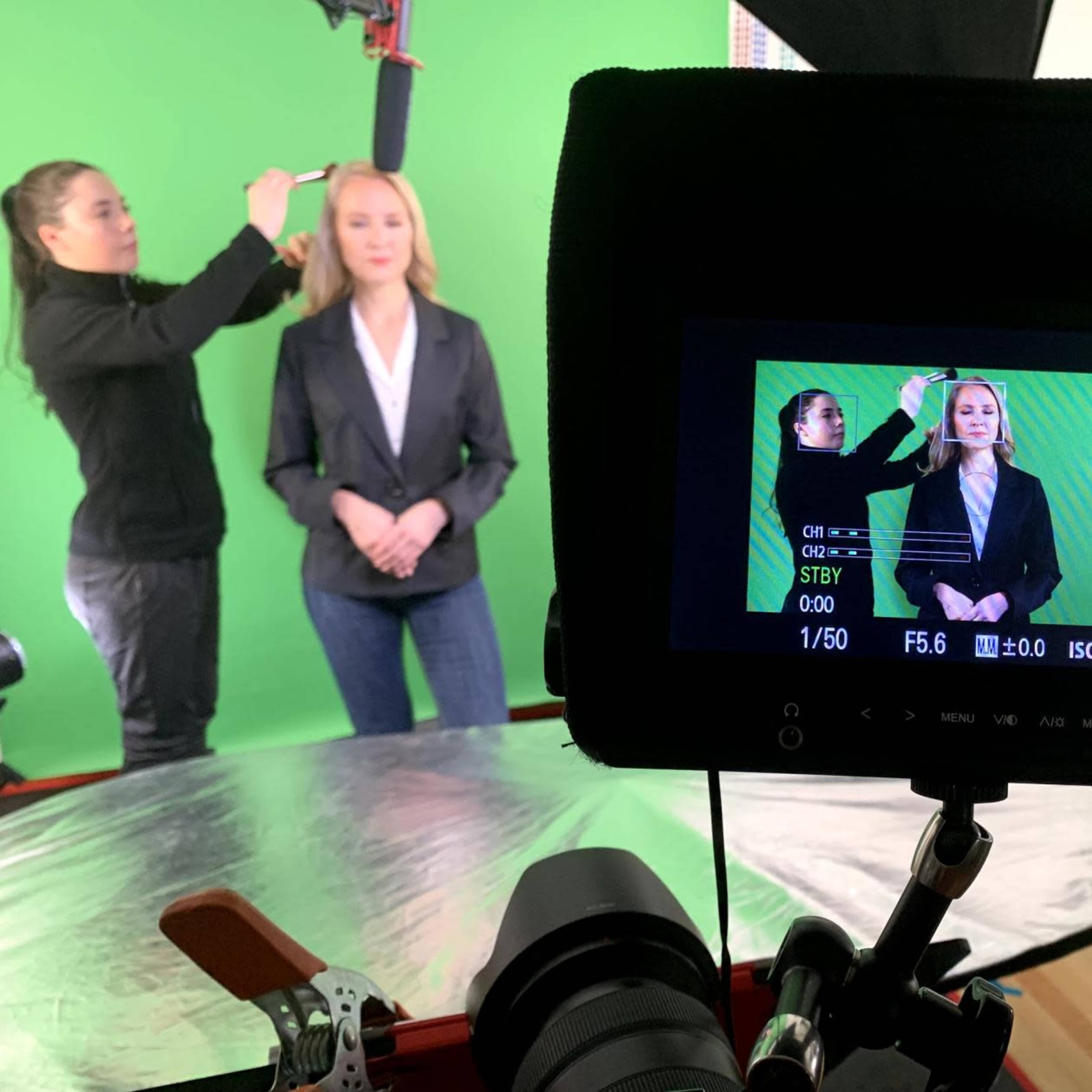 A woman is getting her makeup done in front of a green screen while another woman adjusts a camera setup.