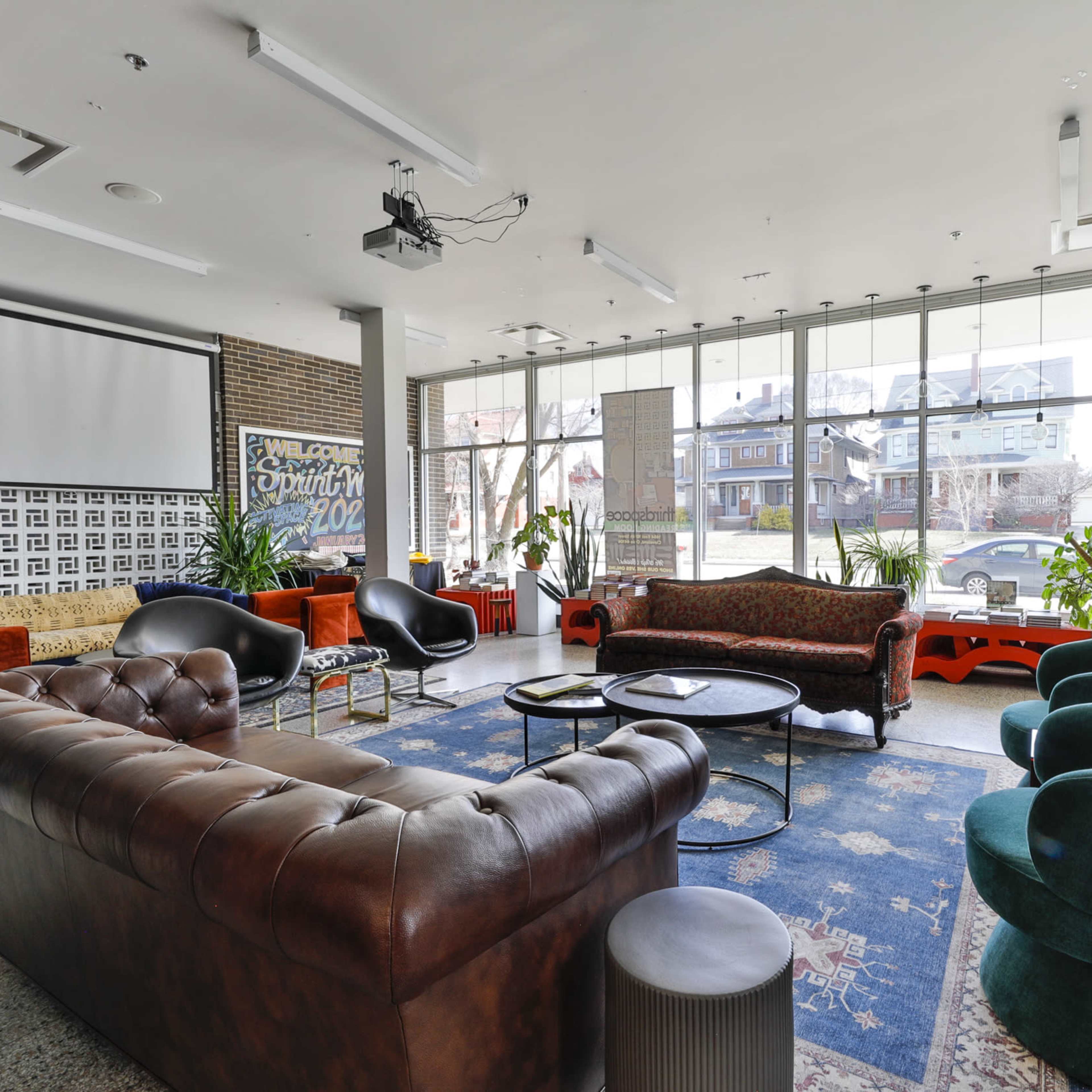 A modern lounge area with a mix of seating options, large windows allowing natural light, and various plants.