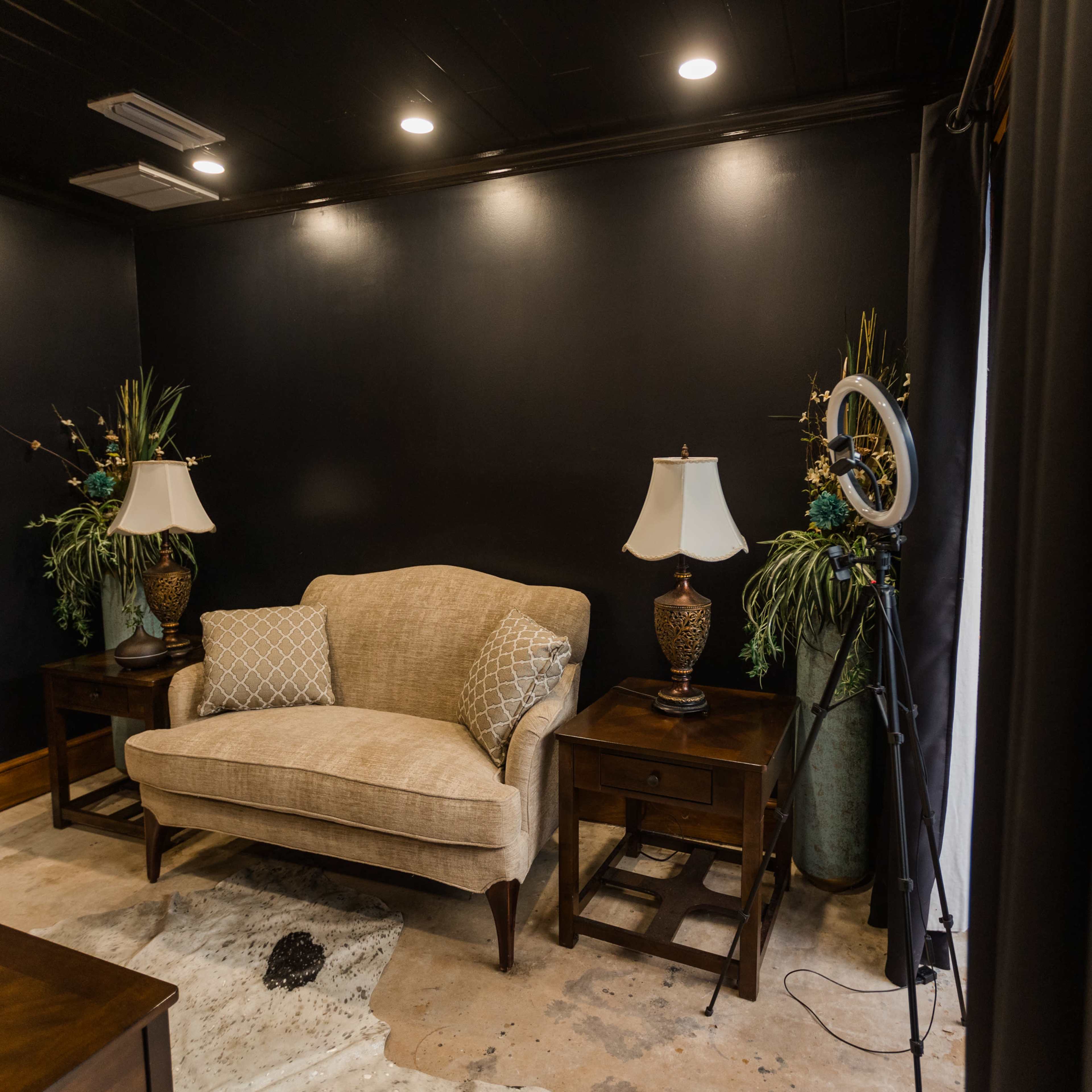 A small room with dark walls, a beige loveseat, two decorative lamps on wooden side tables, and potted plants in the corners.