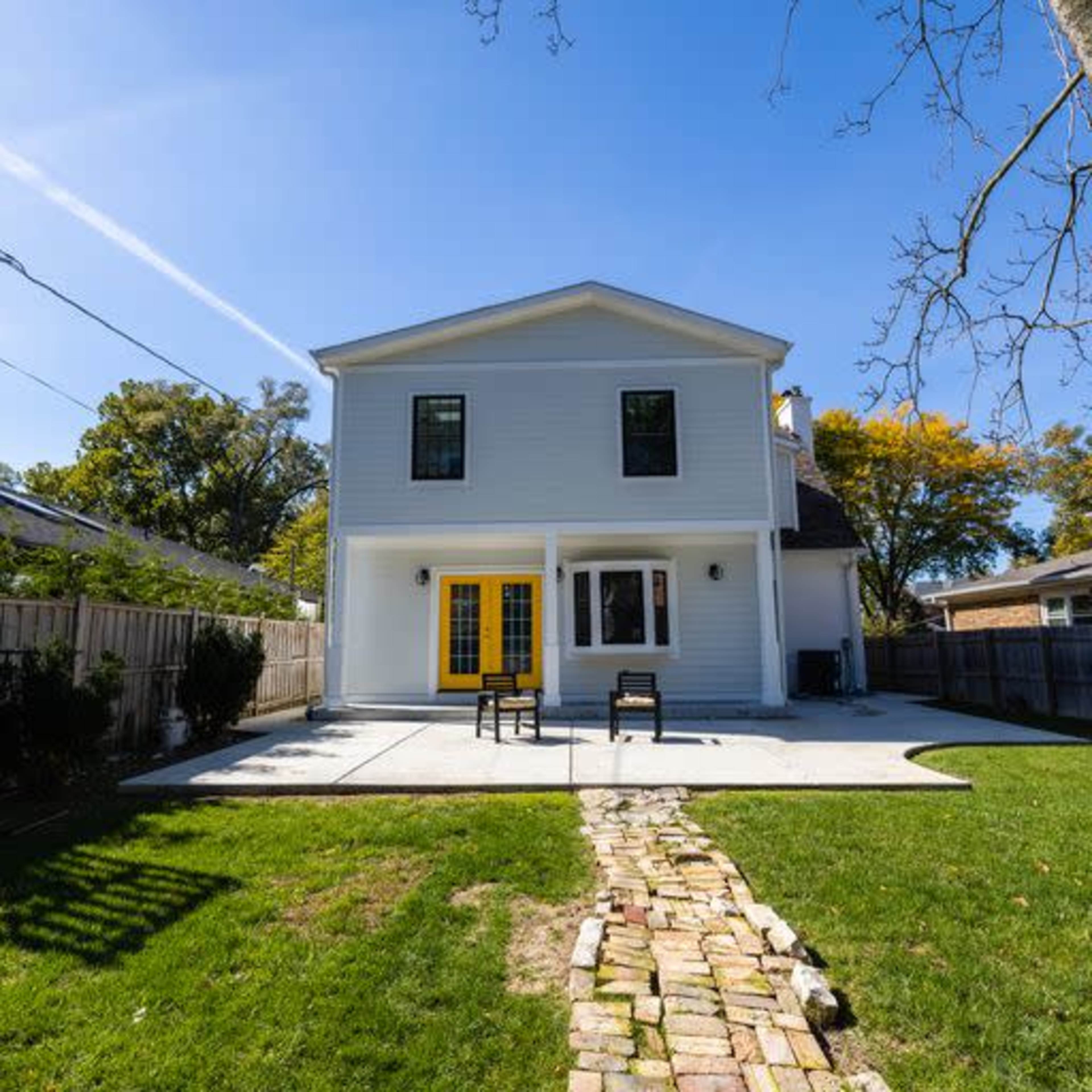 A two-story light blue house with yellow doors sits on a well-maintained lawn, featuring a stone path and patio furniture in the backyard.