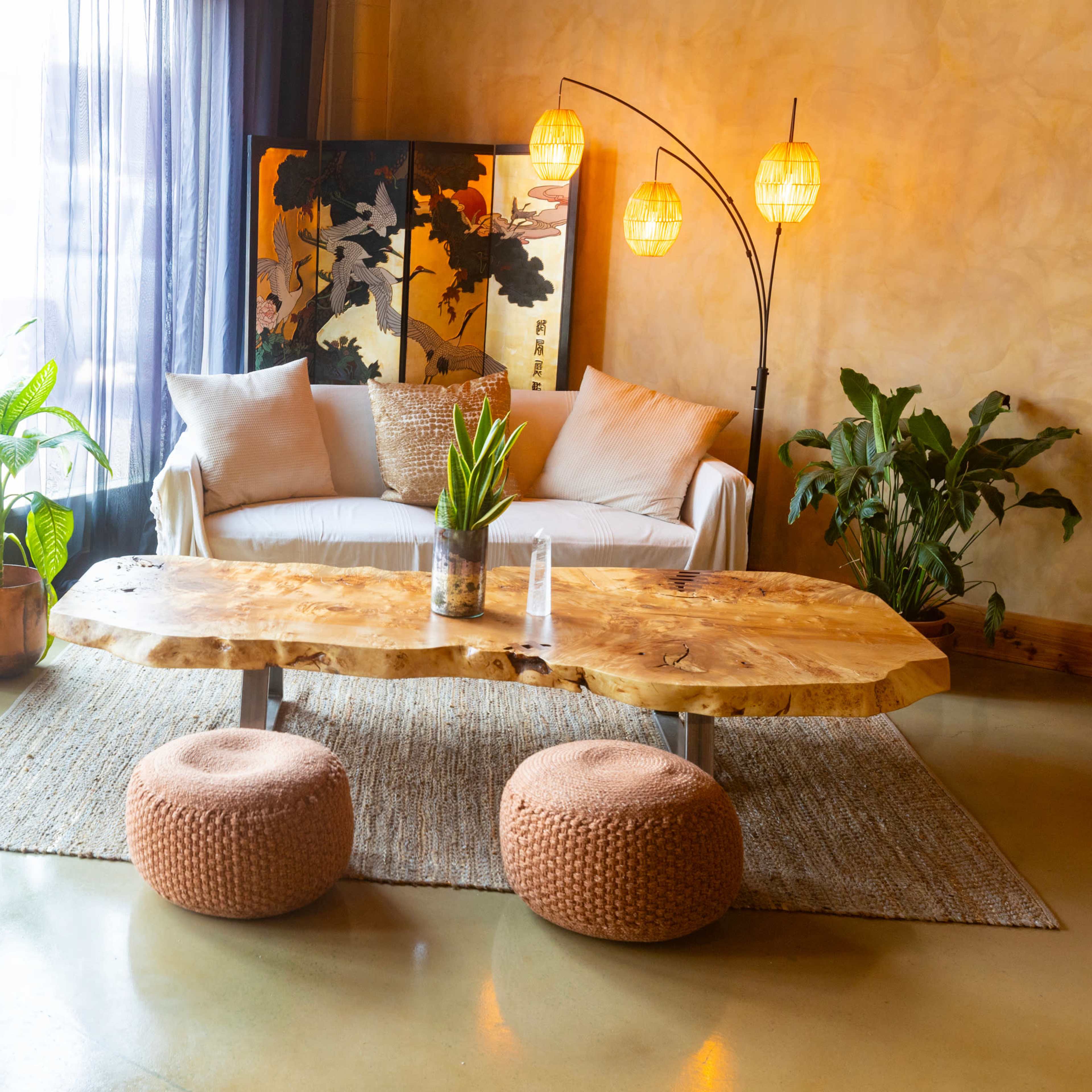 The scene depicts a modern living room with a white sofa, a large wooden coffee table, and various plants, illuminated by natural light filtering through sheer curtains.