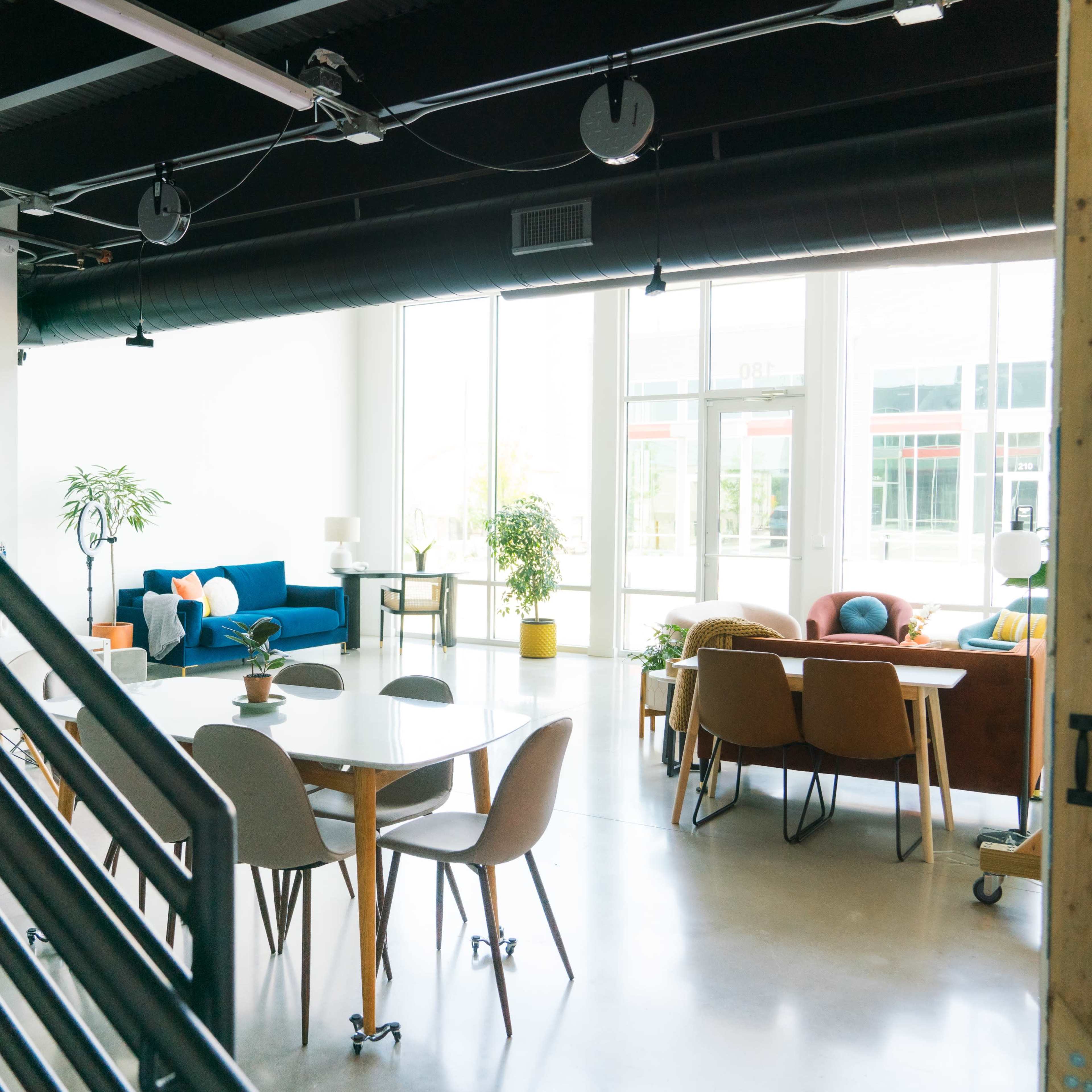 A modern, open-plan workspace features a dining table surrounded by chairs, a staircase, and various seating areas with plants and natural light.