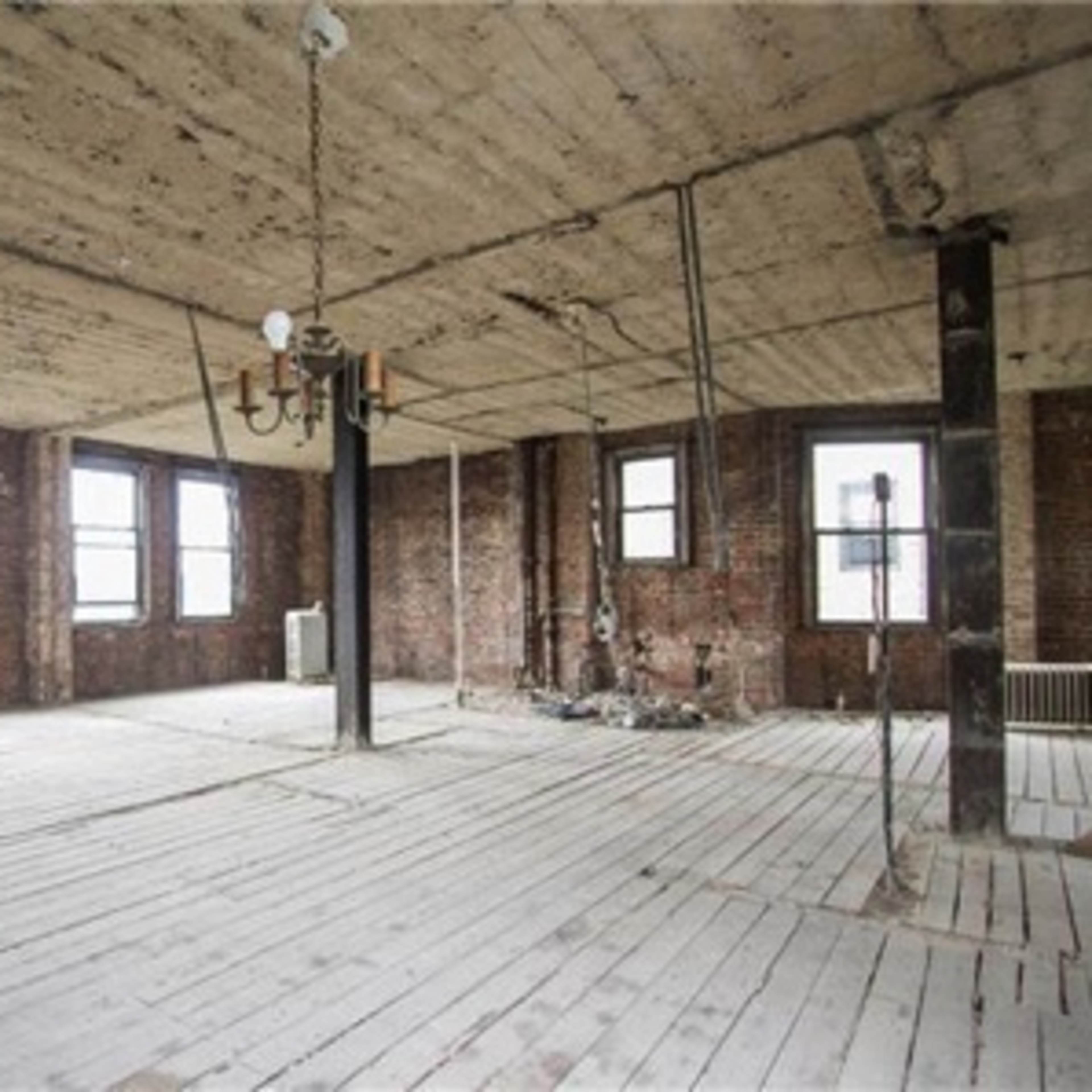An empty, unfinished interior space with exposed brick walls, large windows, and wooden flooring.