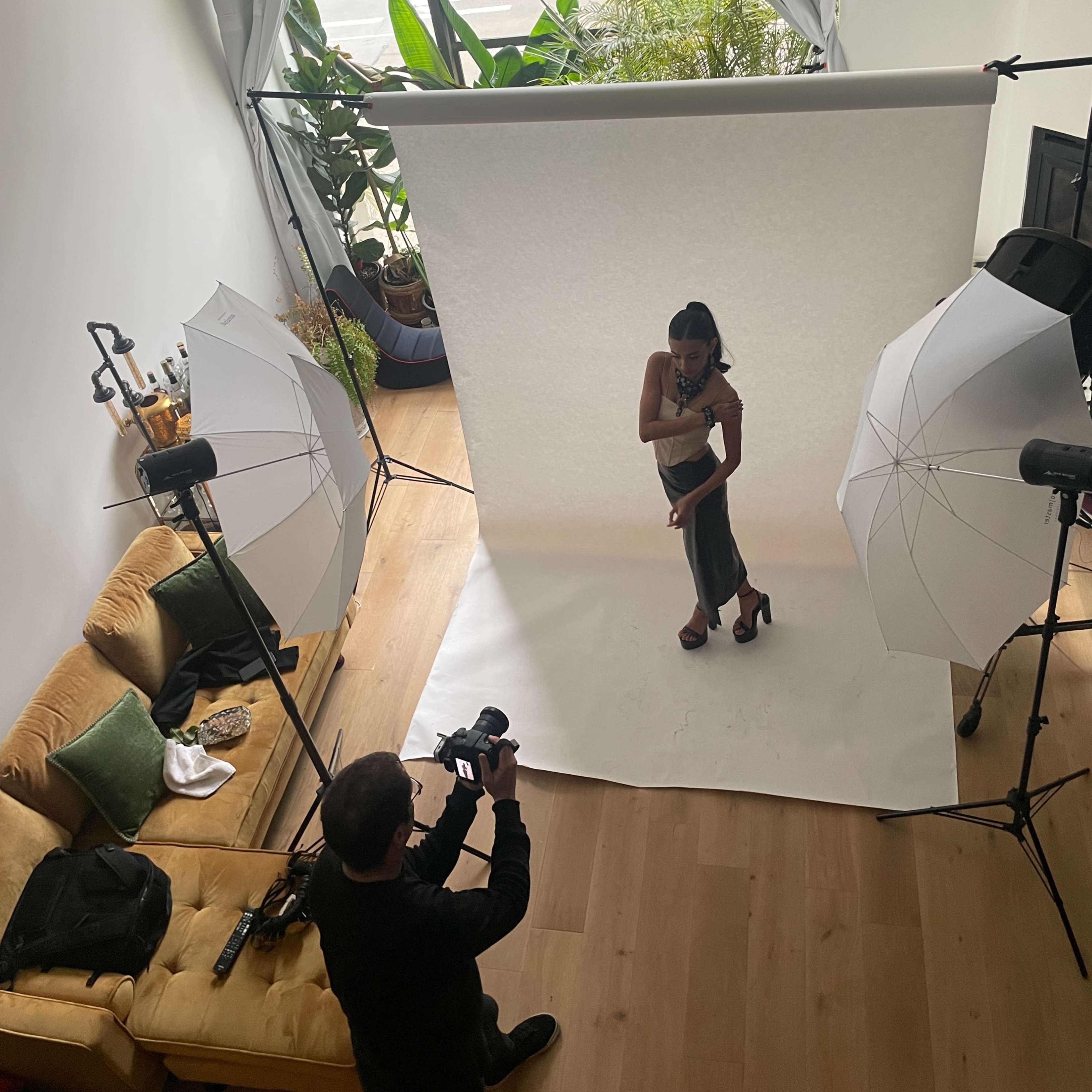 A photographer captures a model's pose in a well-lit indoor space with multiple light setups and a white backdrop.