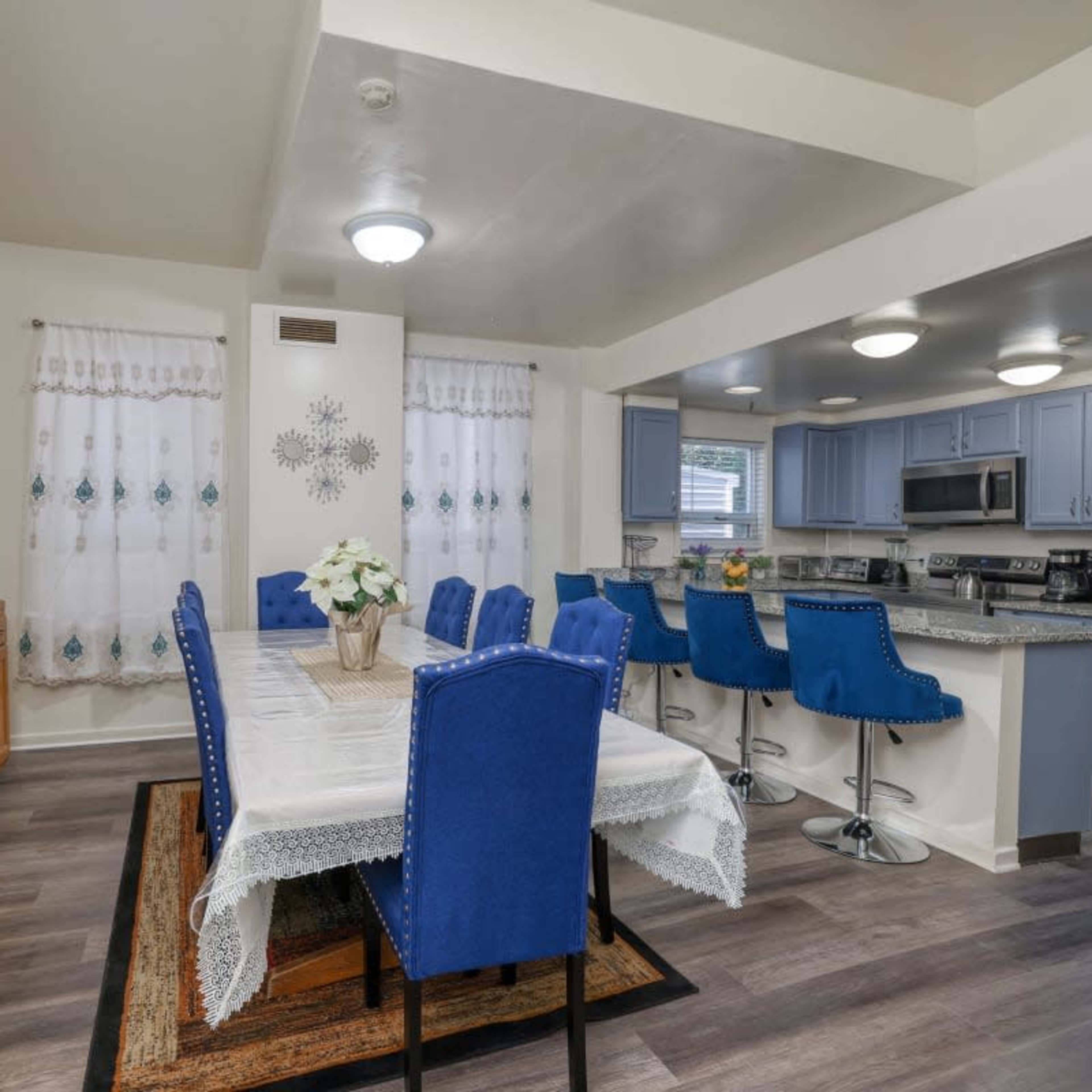 A dining area features a long table surrounded by blue chairs, with a sideboard and an open kitchen in the background.