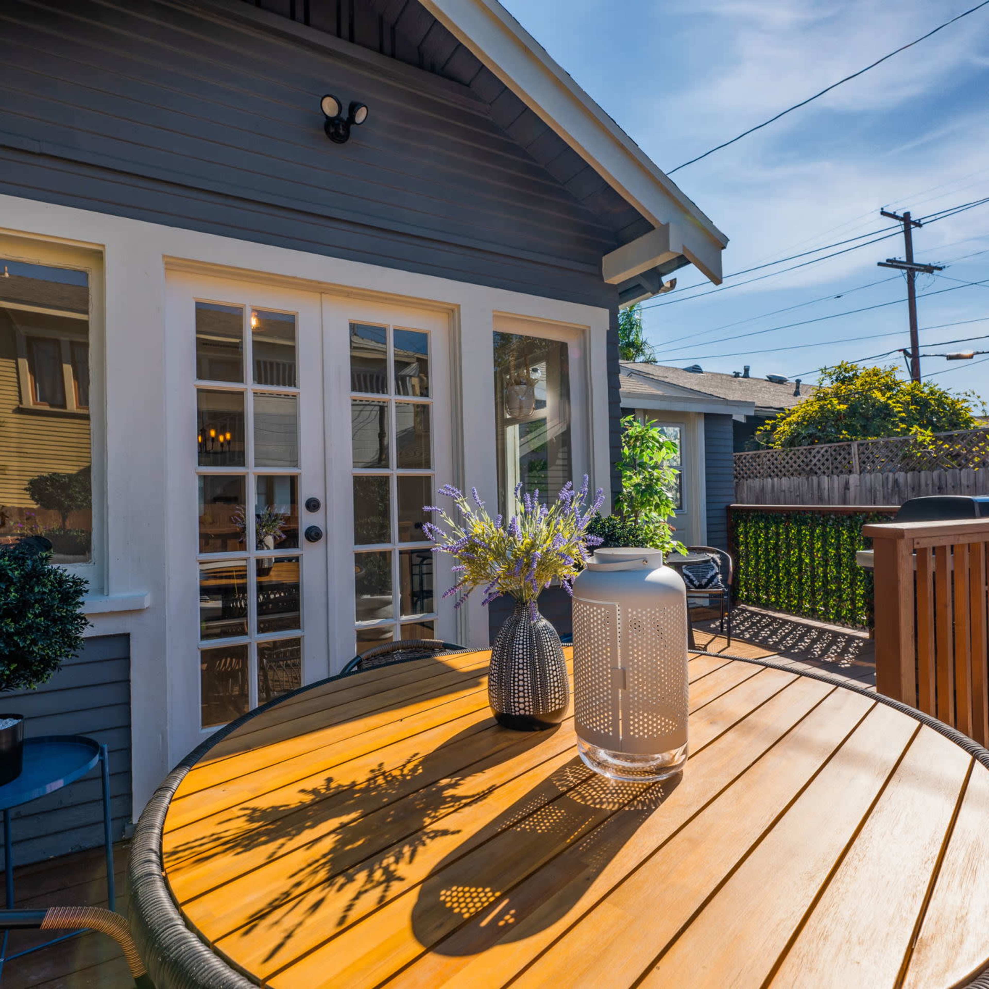 A wooden table with decorative items sits on a patio next to a house with a gray exterior and a view of a fenced backyard.
