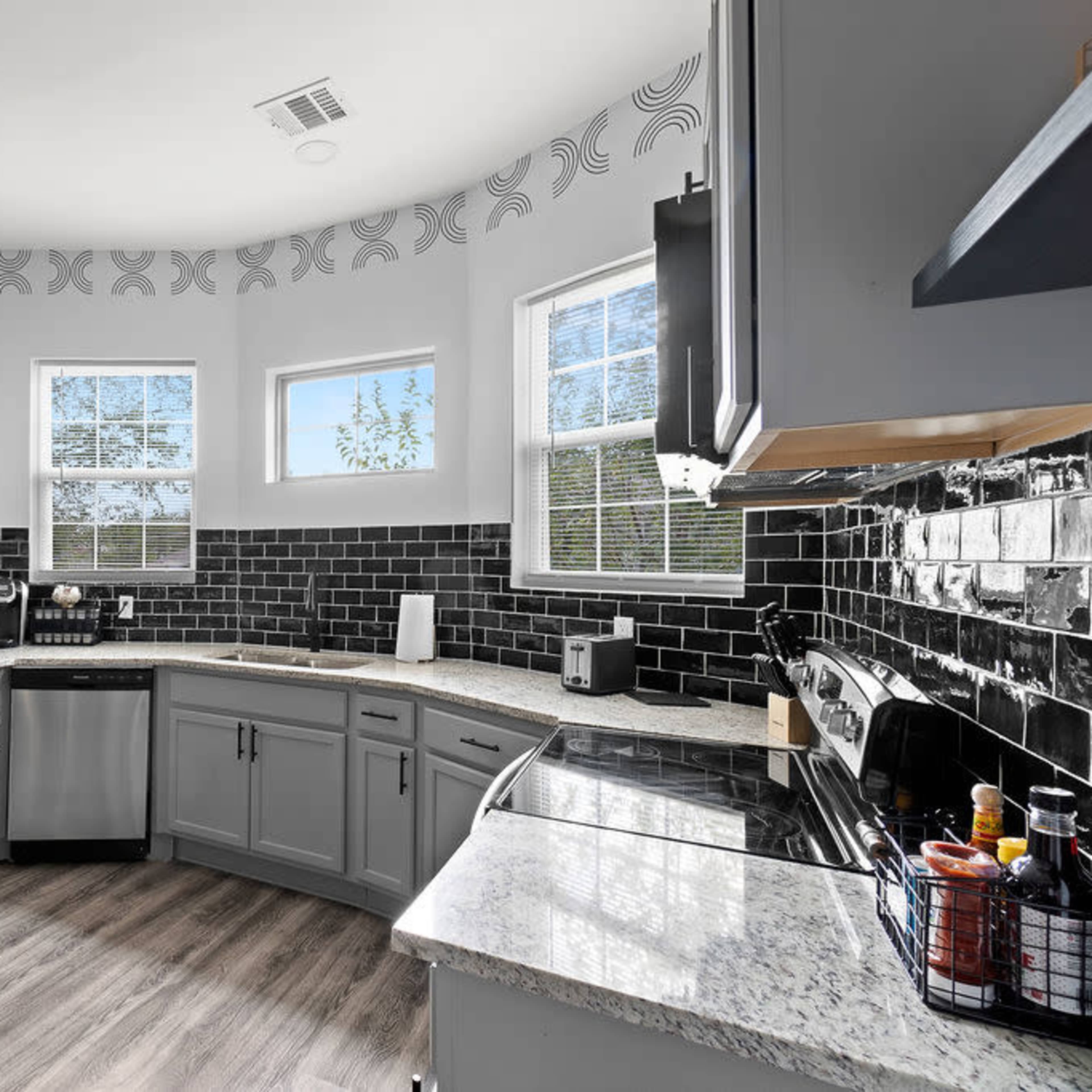 The kitchen features gray cabinets, a black tile backsplash, and three windows providing natural light.