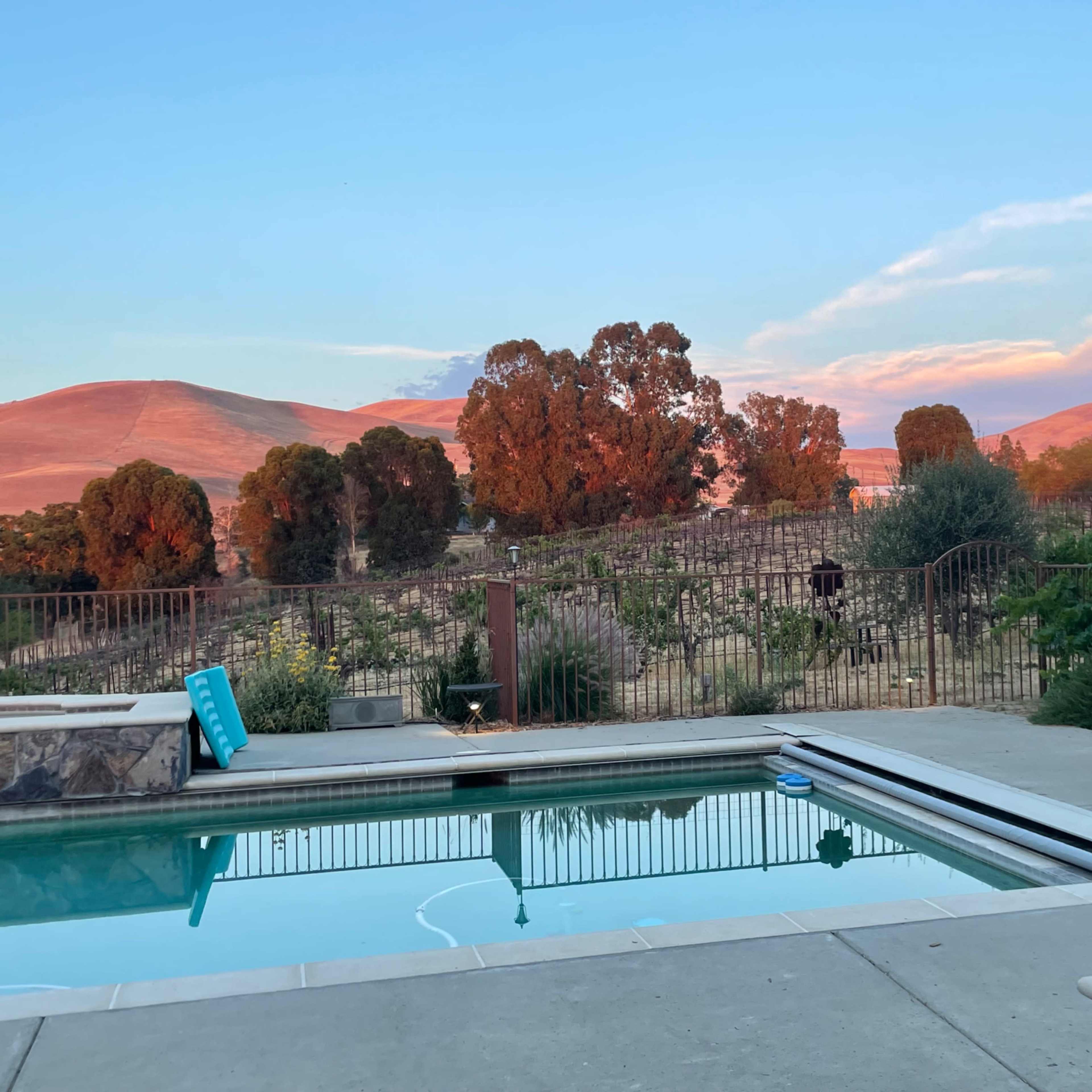 A swimming pool with a clear reflection is surrounded by a metal fence, with rolling hills and trees in the background under a colorful sky.