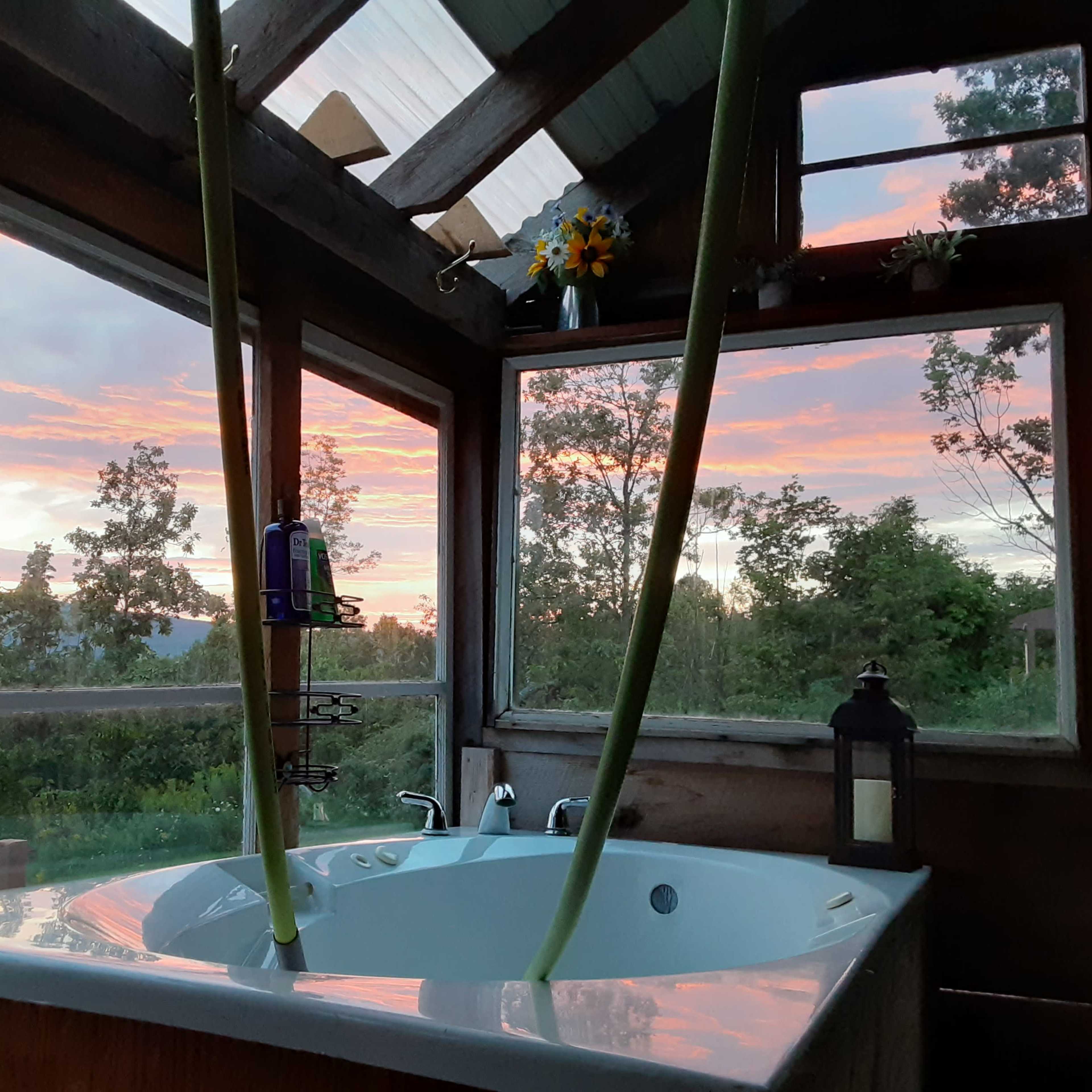 A bathtub inside a wooden structure with large windows, framing a colorful sunset outside.