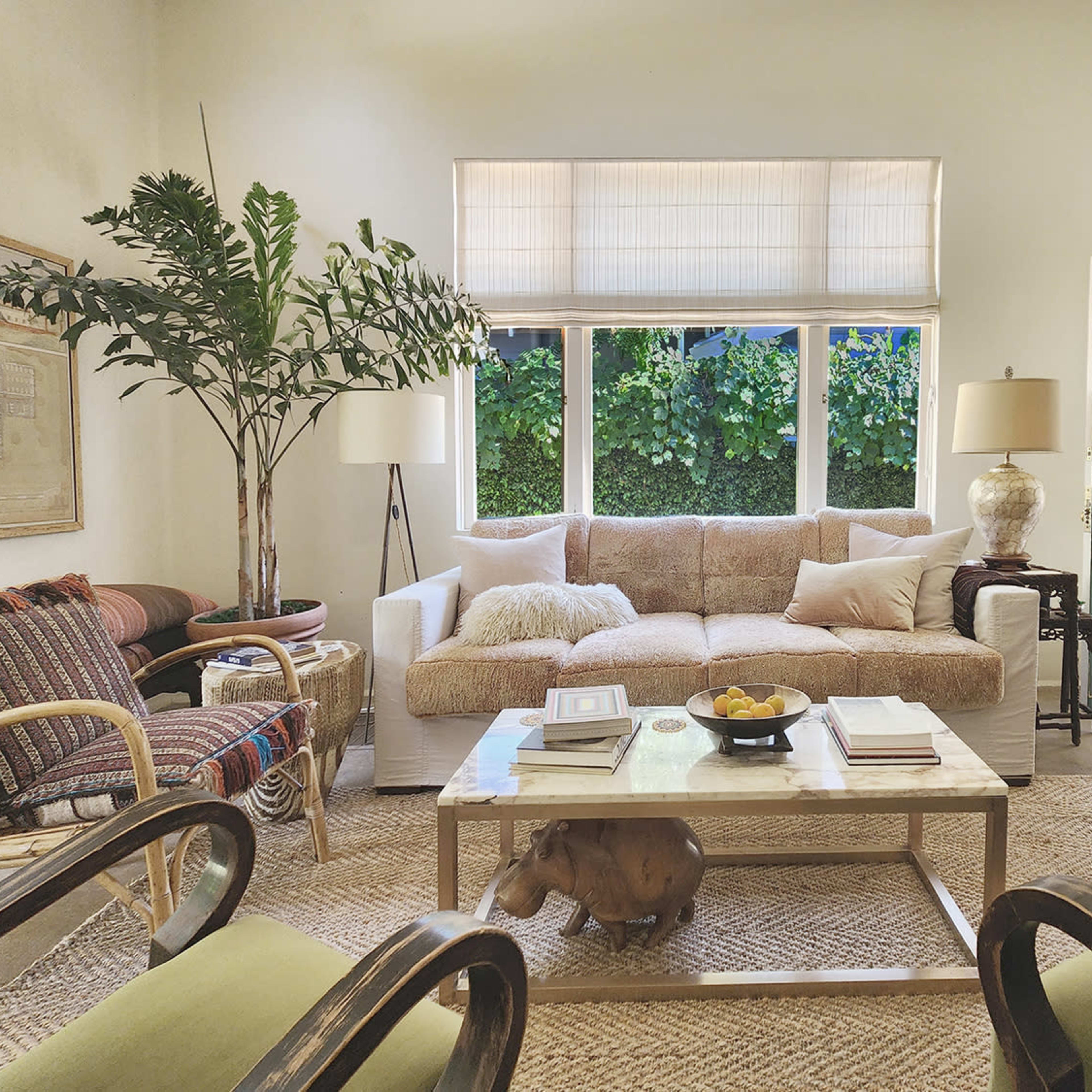 A bright living room features a beige sofa and a coffee table with a bowl of fruit, surrounded by chairs and large windows overlooking greenery.