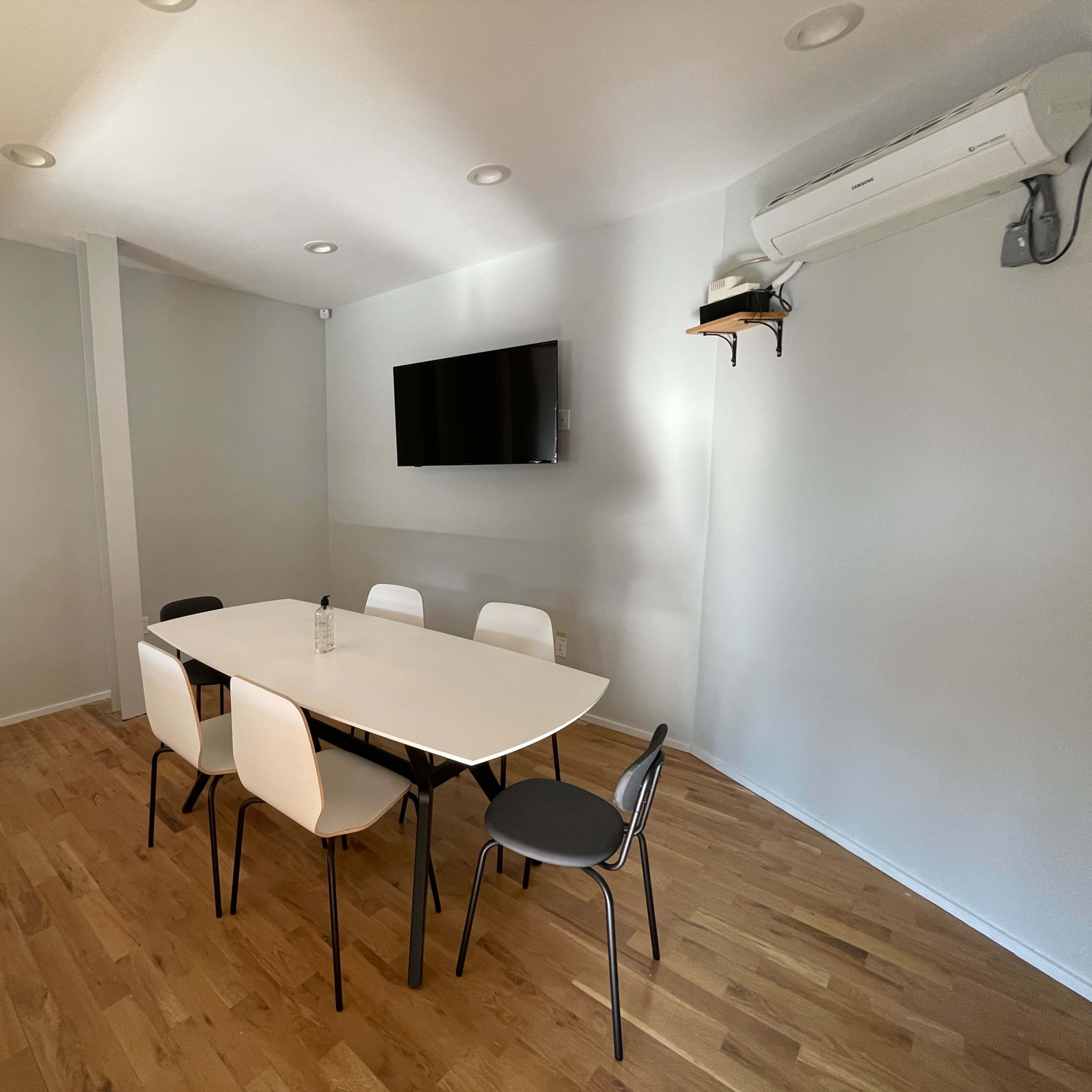 A light-colored meeting room with a large table surrounded by six chairs and a wall-mounted television.