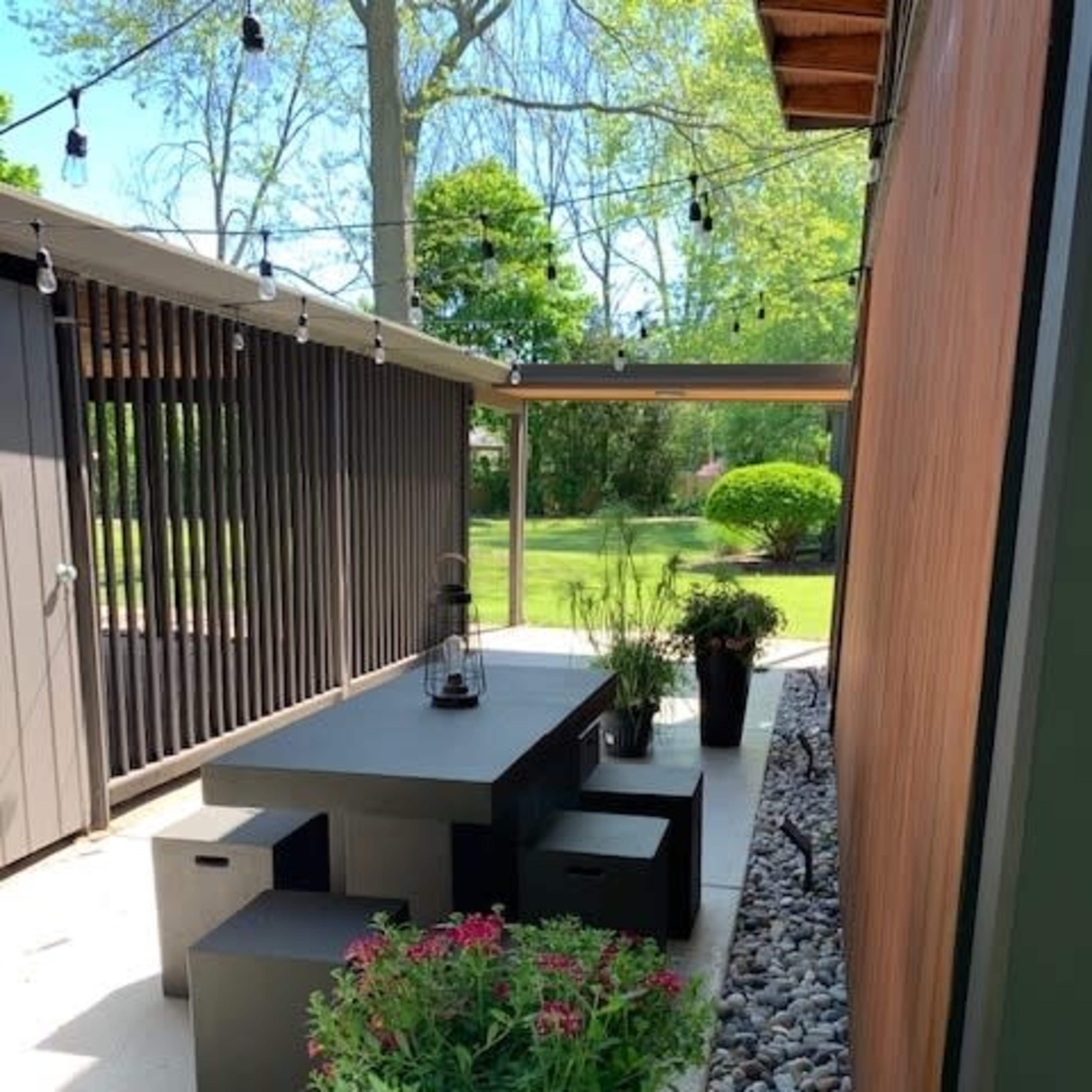 A modern outdoor dining area with a rectangular black table, surrounding square stools, and potted plants, set in a landscaped garden with bright green foliage.