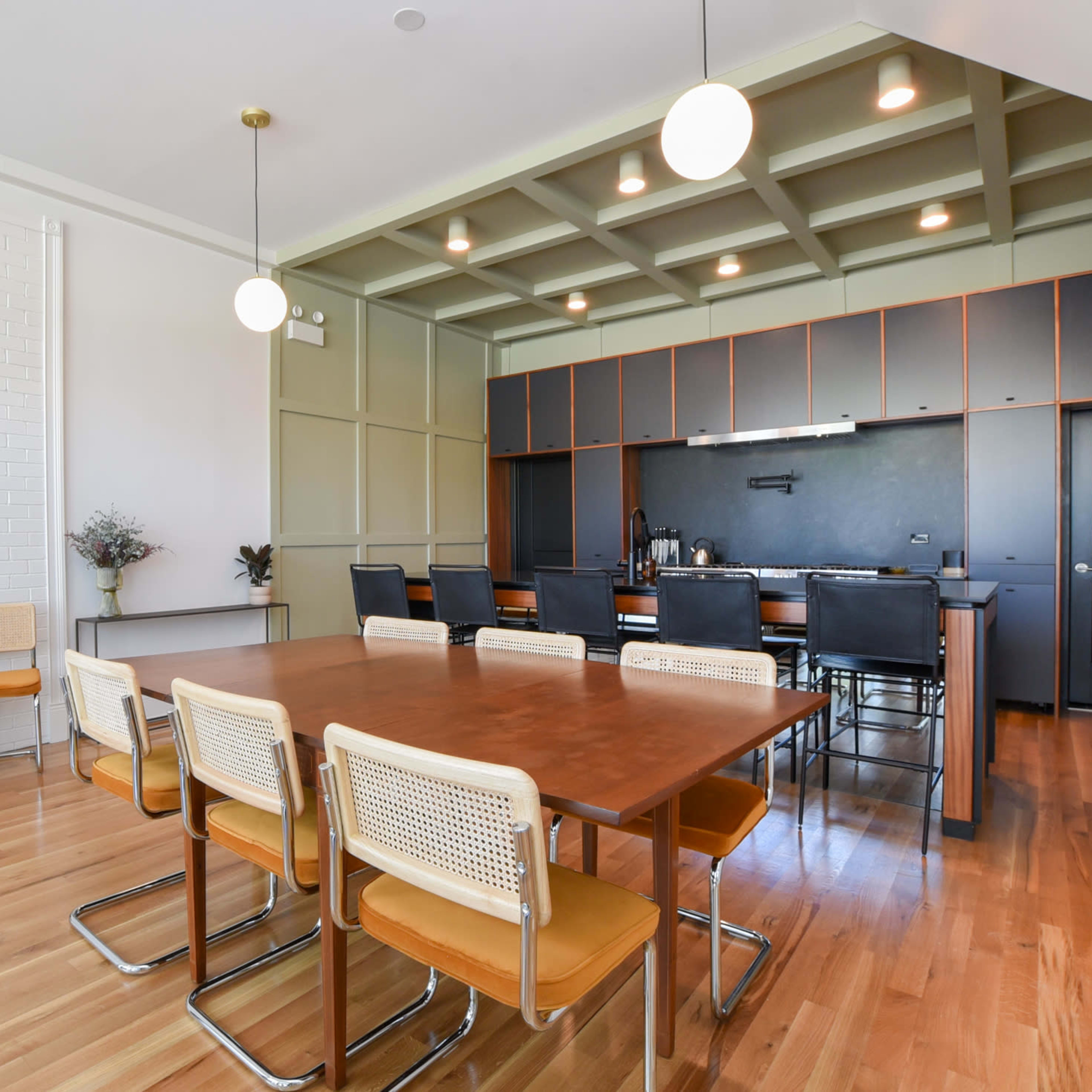 A modern dining area with a wooden table surrounded by metal-framed chairs, adjacent to a kitchen featuring dark cabinetry and overhead lighting.