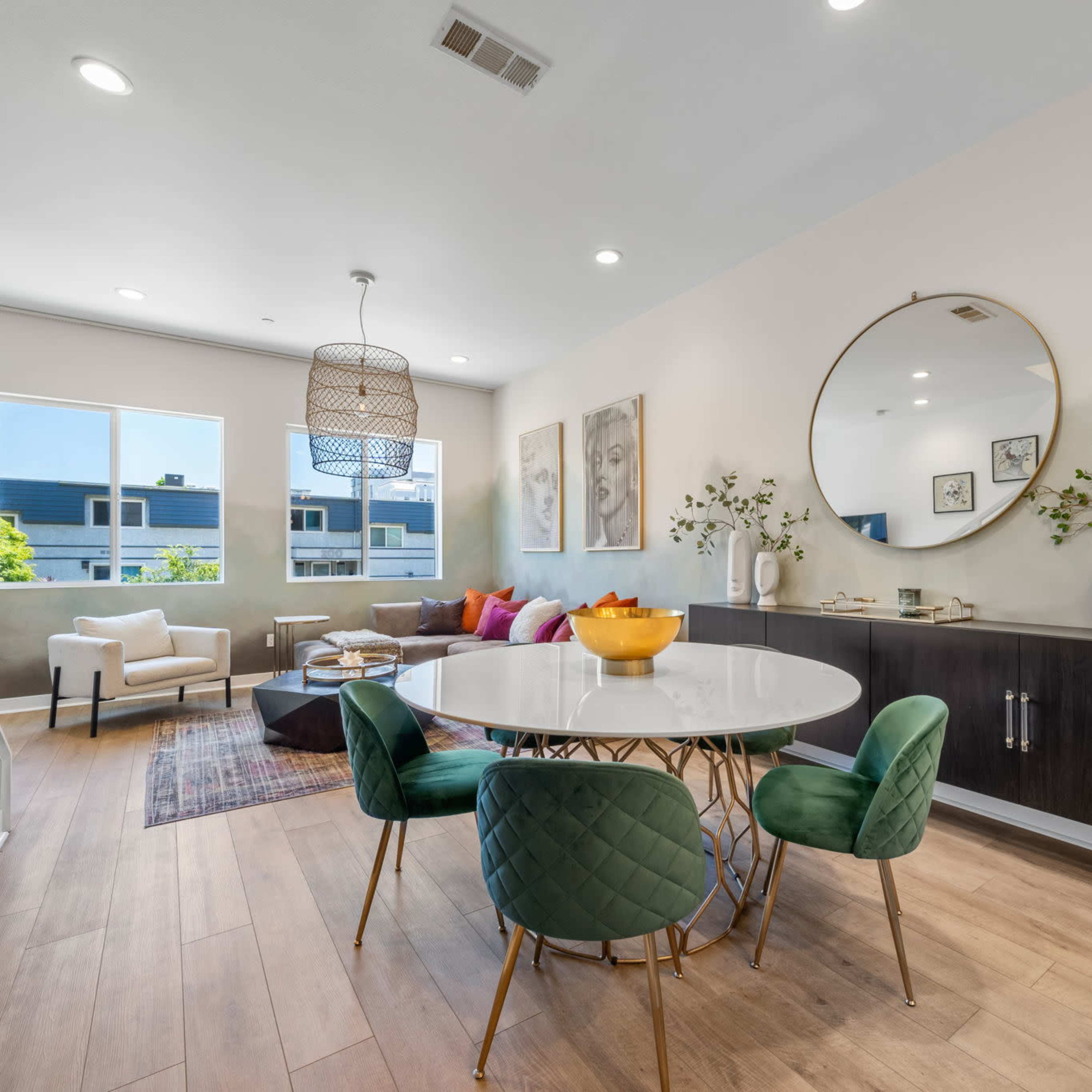 A modern dining area features a round table with green chairs, surrounded by a spacious living space with a sofa, decorative pillows, and large windows.