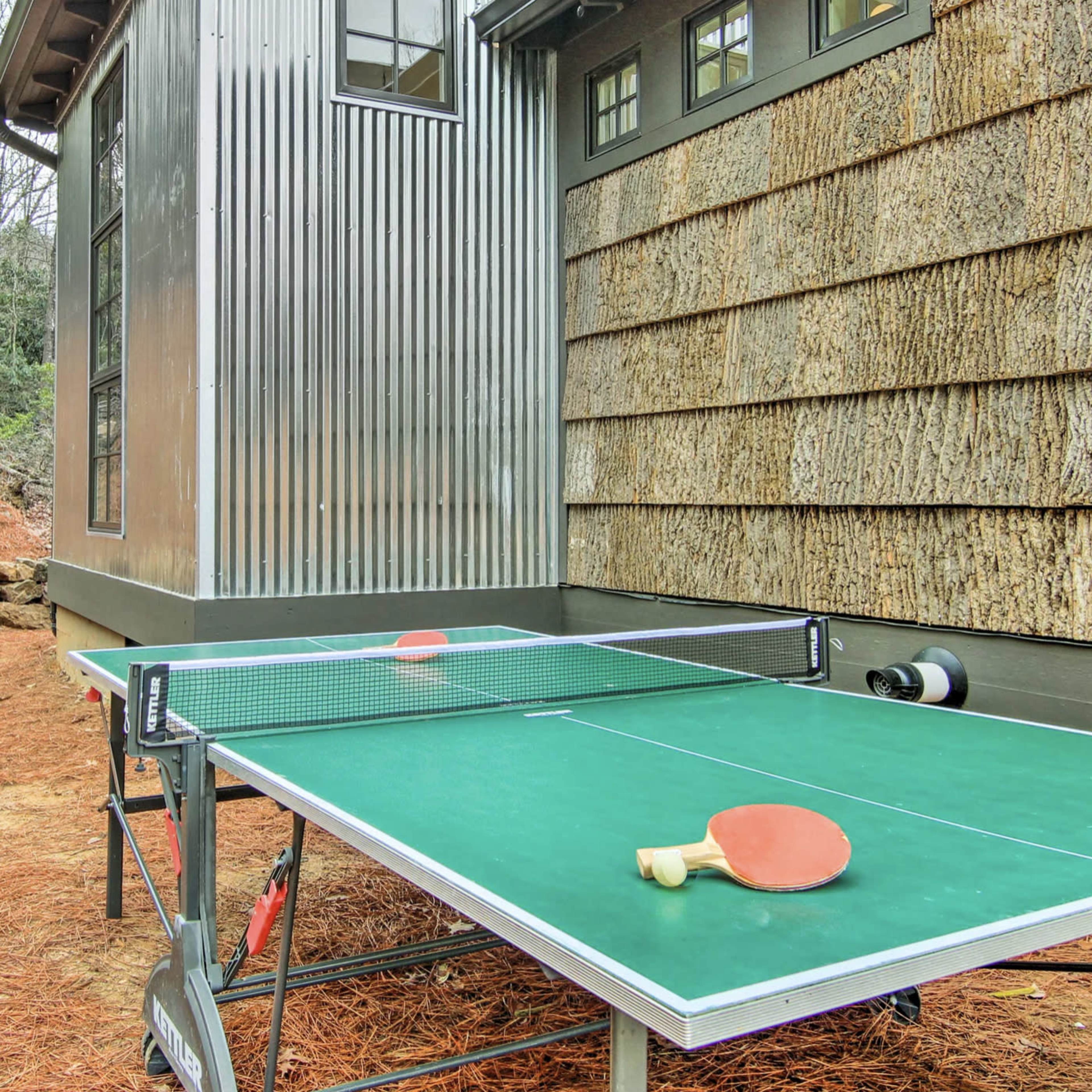 A ping pong table with paddles sits outside next to a building with corrugated metal and wooden siding.