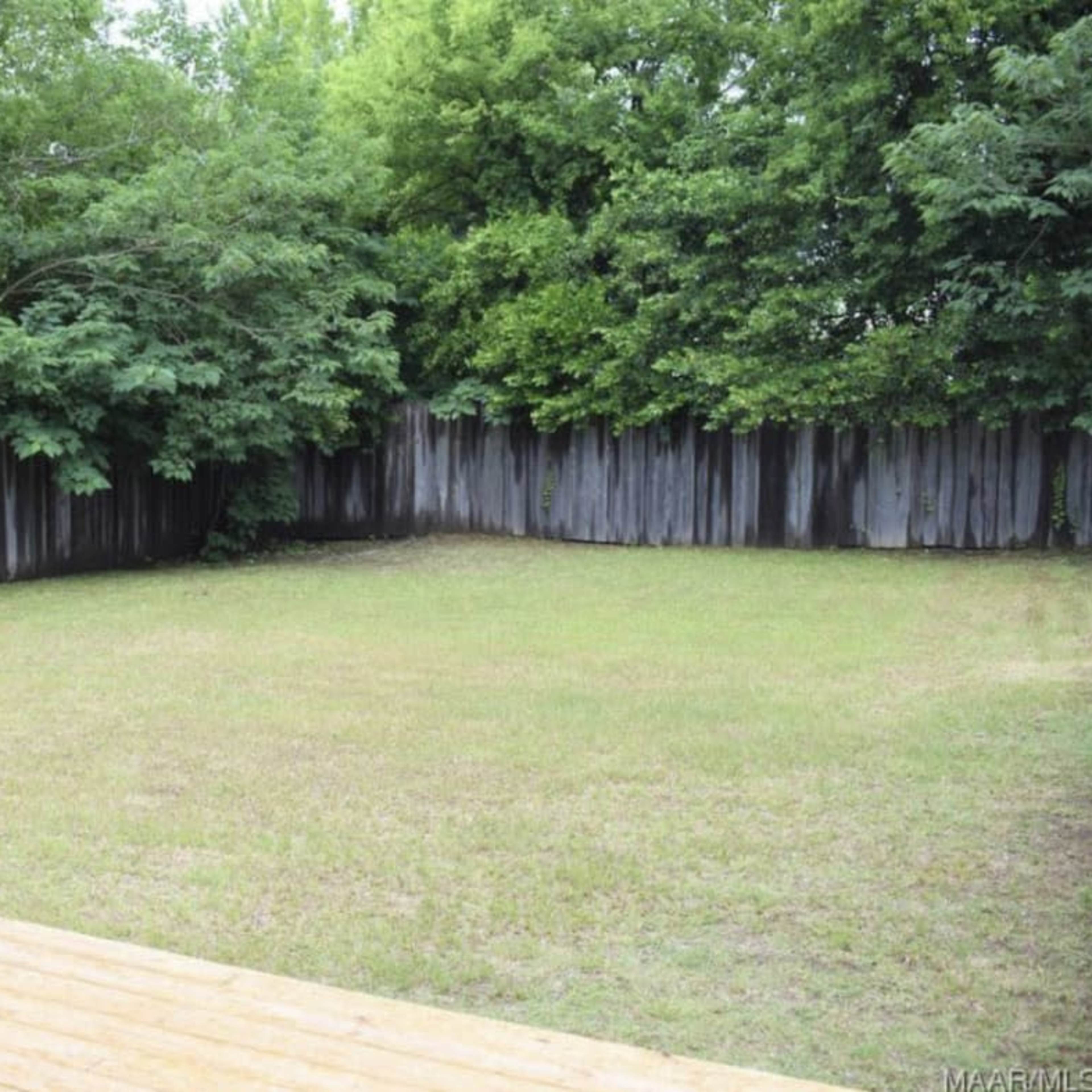 A grassy backyard is enclosed by a wooden fence and surrounded by trees.