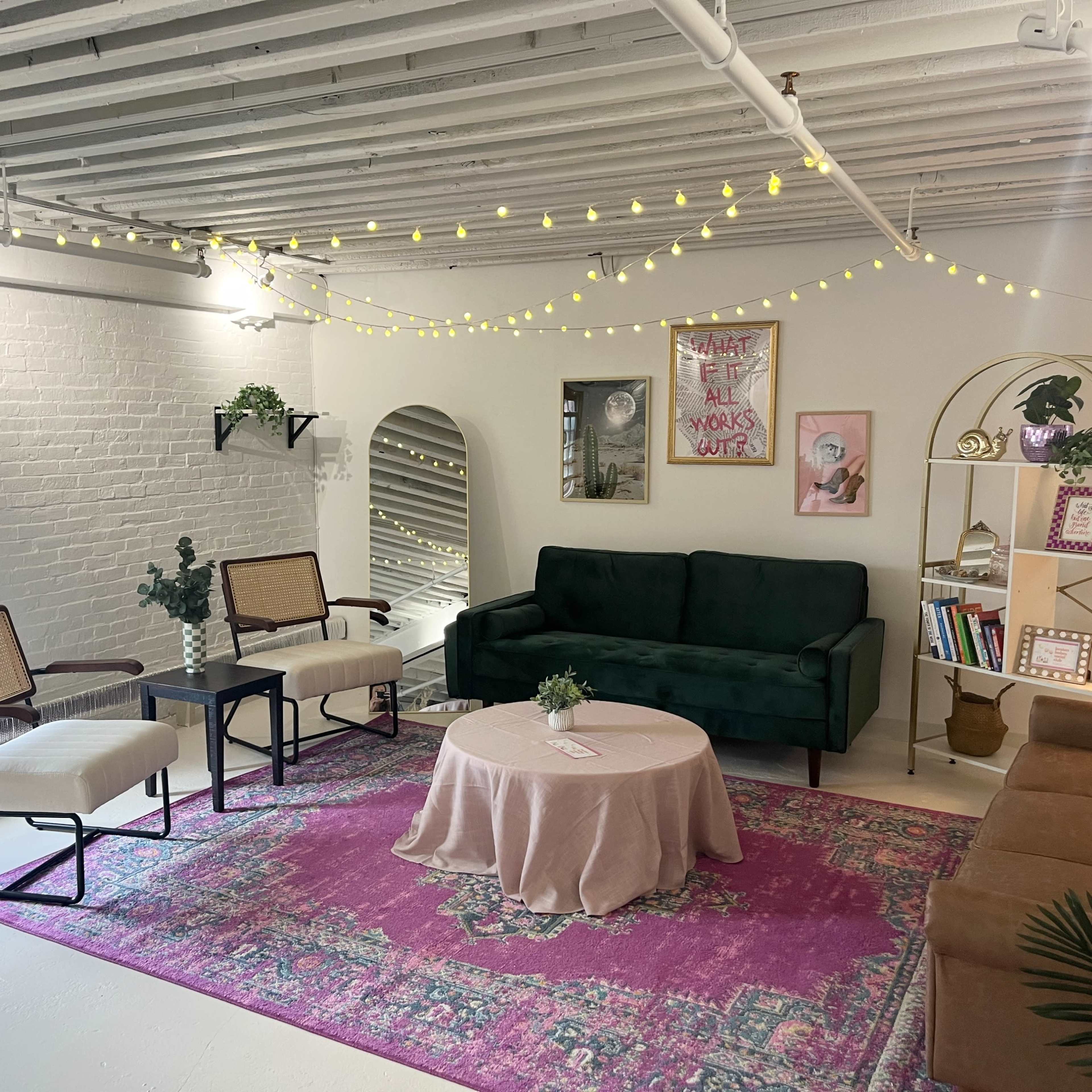 A modern living space with a green sofa, two chairs, a round coffee table on a pink rug, and a bookshelf surrounded by warm lighting.