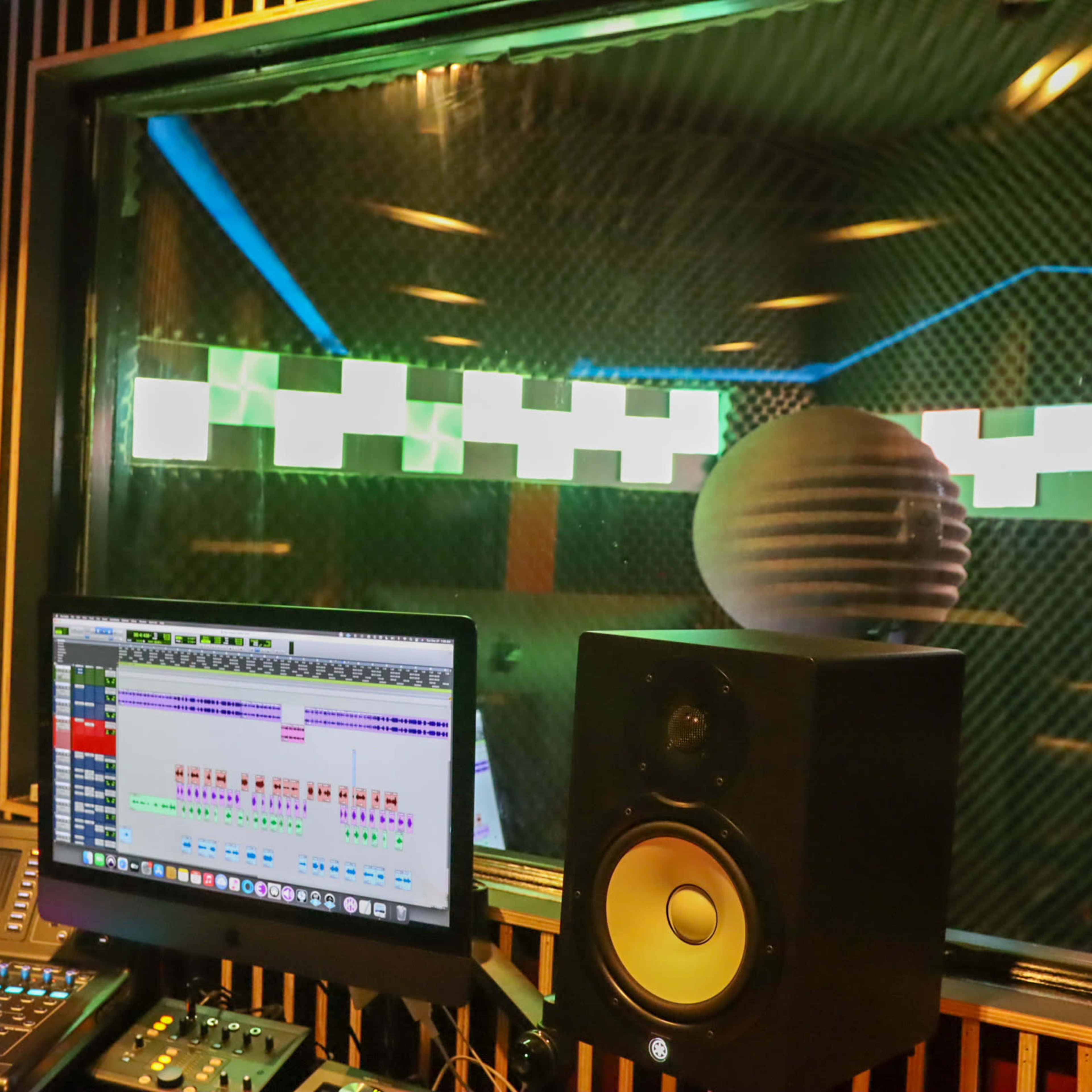 A recording studio control room features a computer display with audio editing software and a speaker, with a soundproof window showing the recording area behind it.