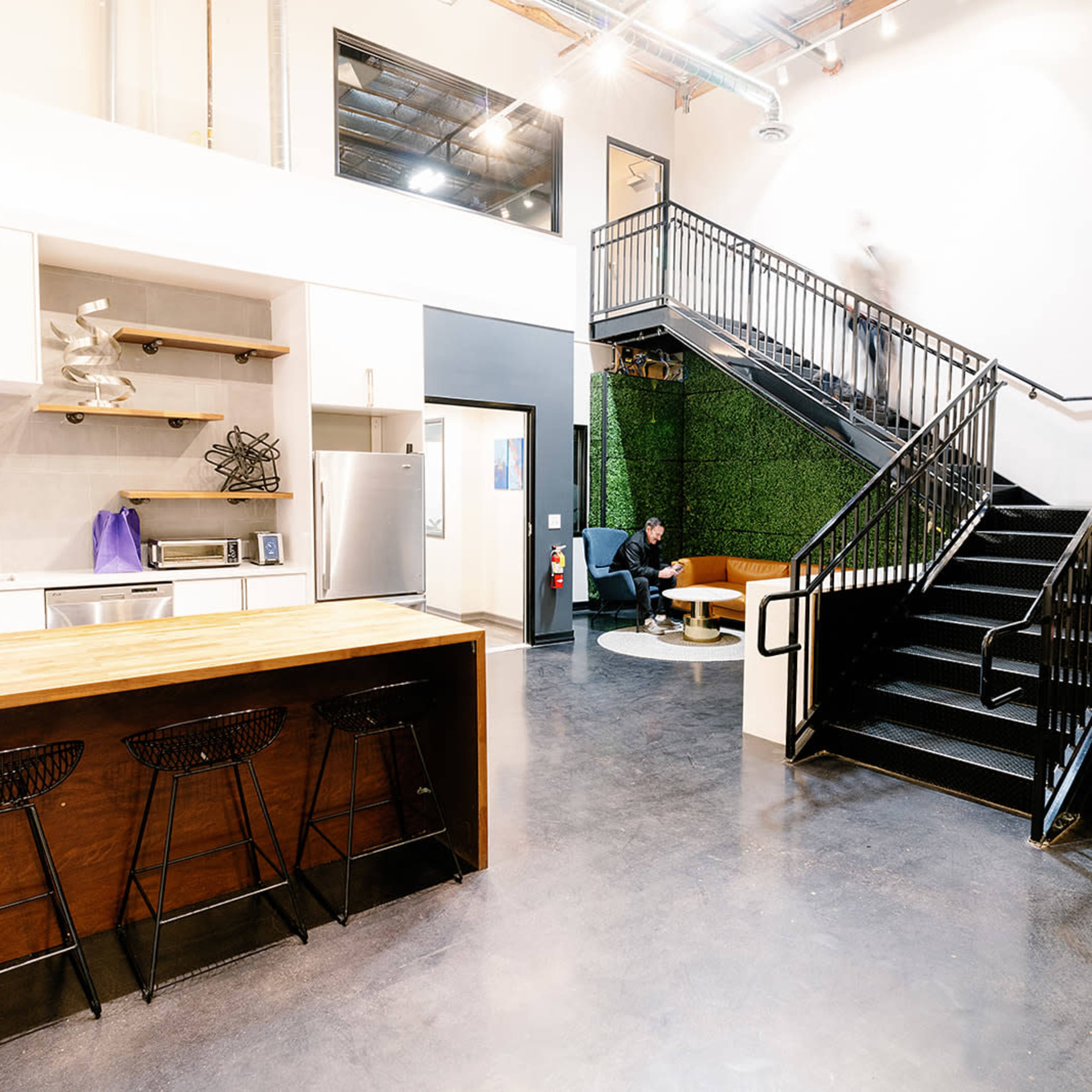 A modern open-concept workspace with a kitchen area, a staircase, and green wall accents, featuring a person sitting in a chair near the staircase.