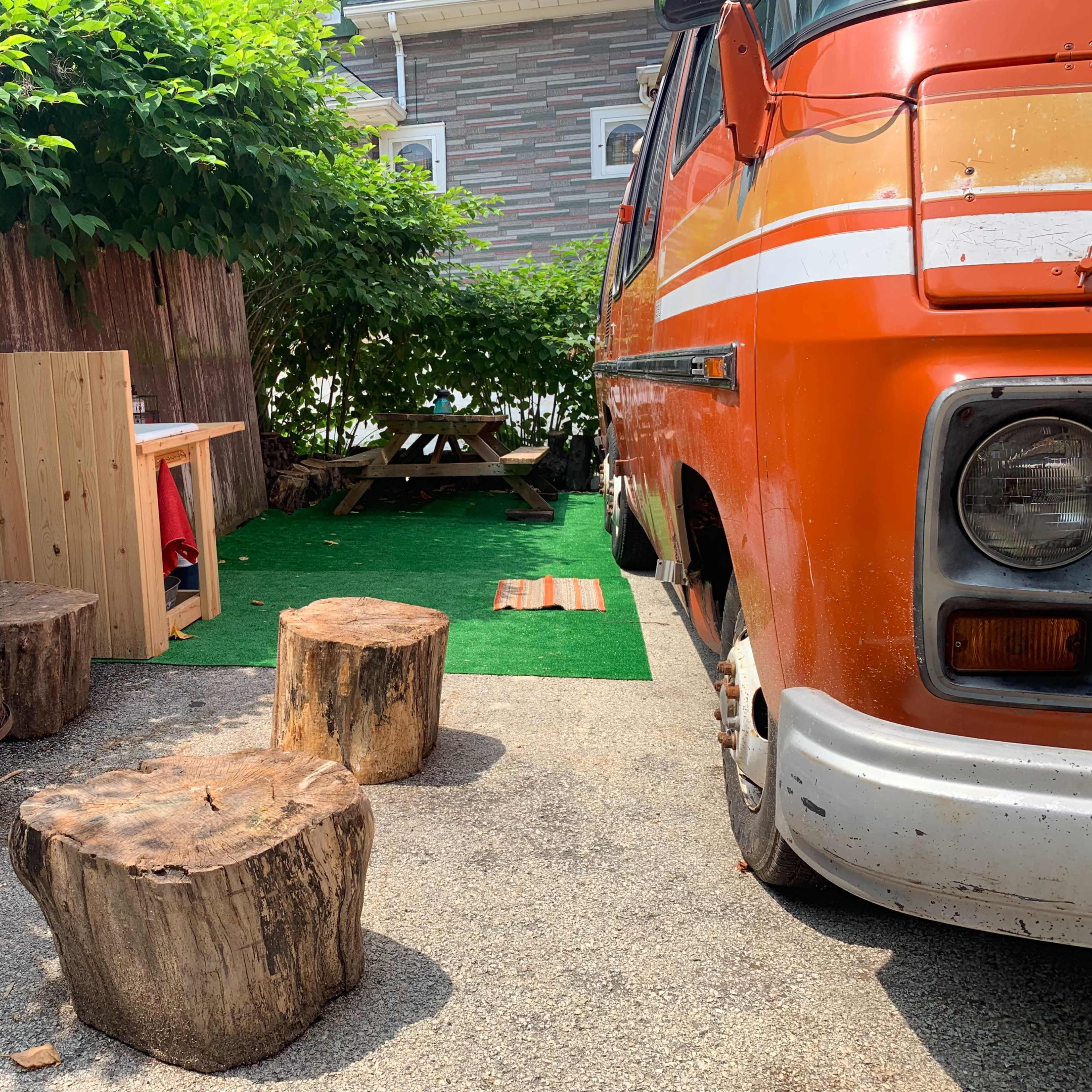 An orange van is parked next to a small outdoor seating area with wooden stools and a picnic table, surrounded by greenery and a stone path.