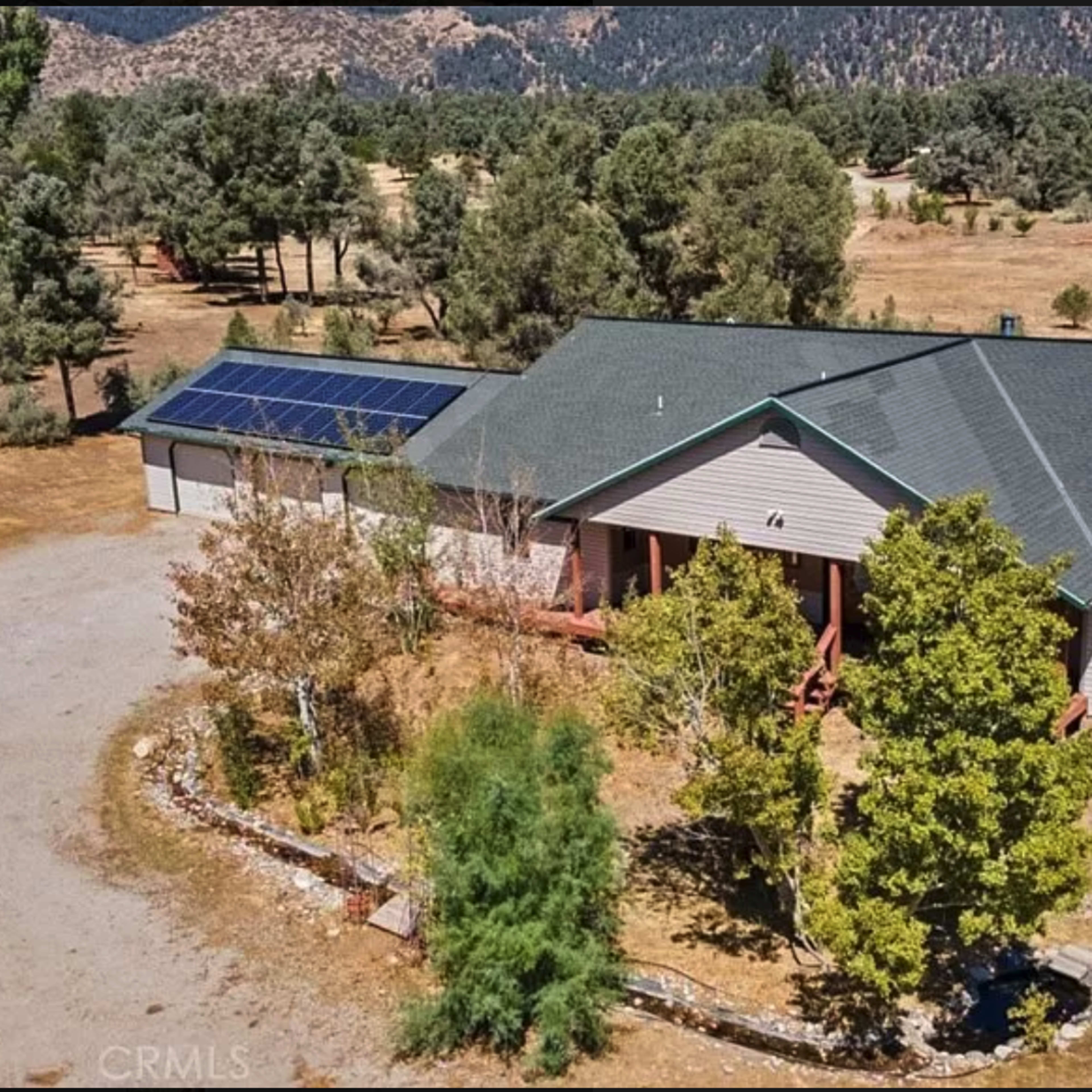 A single-story house with a solar panel on the roof, surrounded by trees and a large open area.